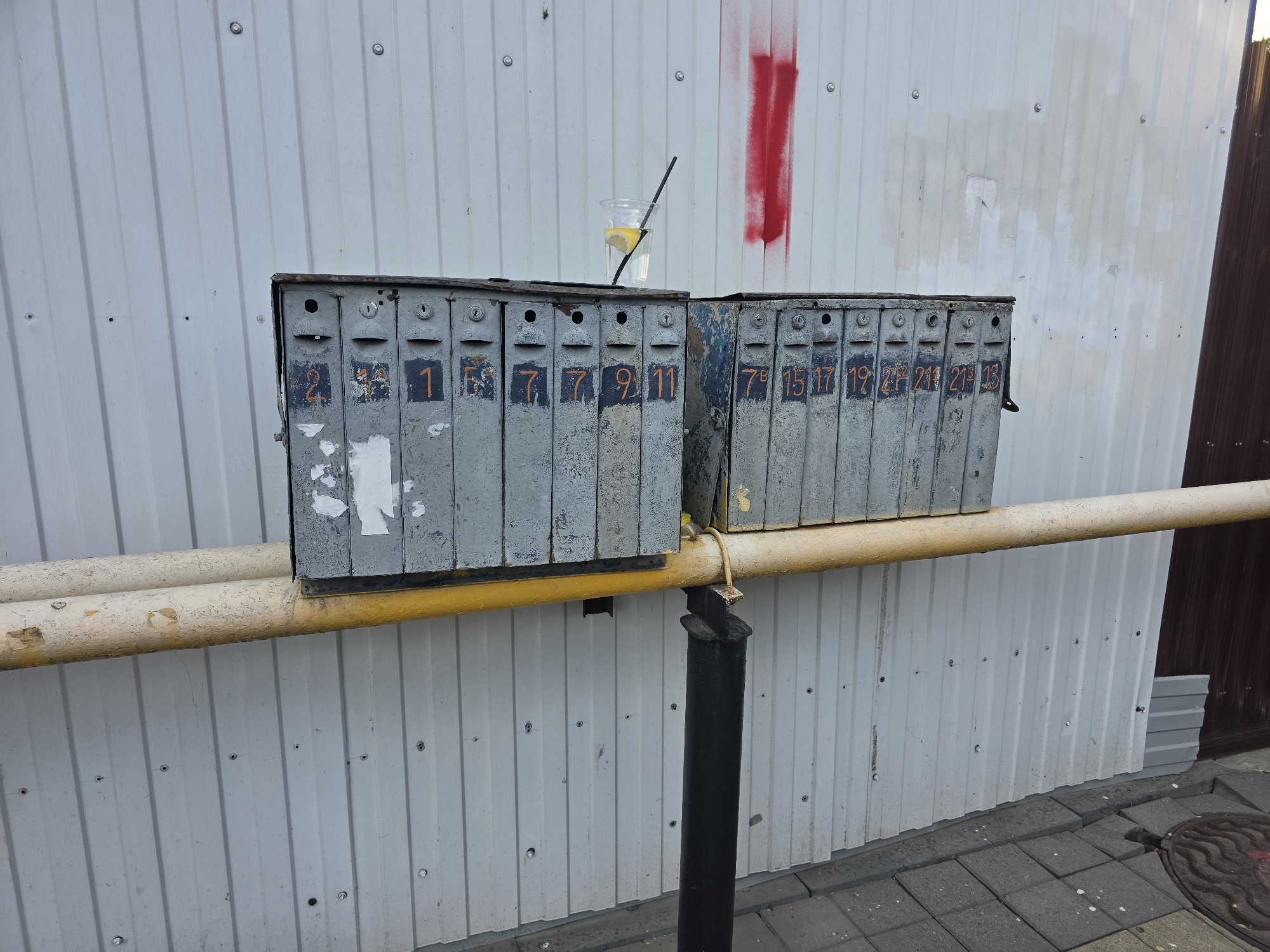 The image depicts a row of old, weathered metal mailboxes mounted on a light-colored corrugated metal wall. The mailboxes are gray and show signs of wear and tear, with some peeling paint and visible rust. Each mailbox is numbered. On the left, the numbers visible are 1, 5, 7, 7, 9, and 11. On the right, the numbers visible are 7B, 15, 17, 19, 2P, 21, and 13. There are two clusters of mailboxes, separated by a gap. A yellow pipe runs horizontally in front of the mailboxes, supported by a black metal post.
On top of the right cluster of mailboxes, there is a clear plastic cup containing a yellow lemon slice and a black straw, filled with a clear liquid. Above the mailboxes, a red vertical spray paint mark is visible on the wall. The wall itself is made of corrugated metal sheets, fastened with visible screws. To the right of the mailboxes, a section of darker brown corrugated metal is visible. The ground in front of the wall is paved with gray interlocking bricks. A circular manhole cover with a patterned surface is partially visible in the lower right corner. The scene appears to be outdoors, likely in an urban or apartment complex setting. The overall impression is one of a functional but neglected outdoor space.