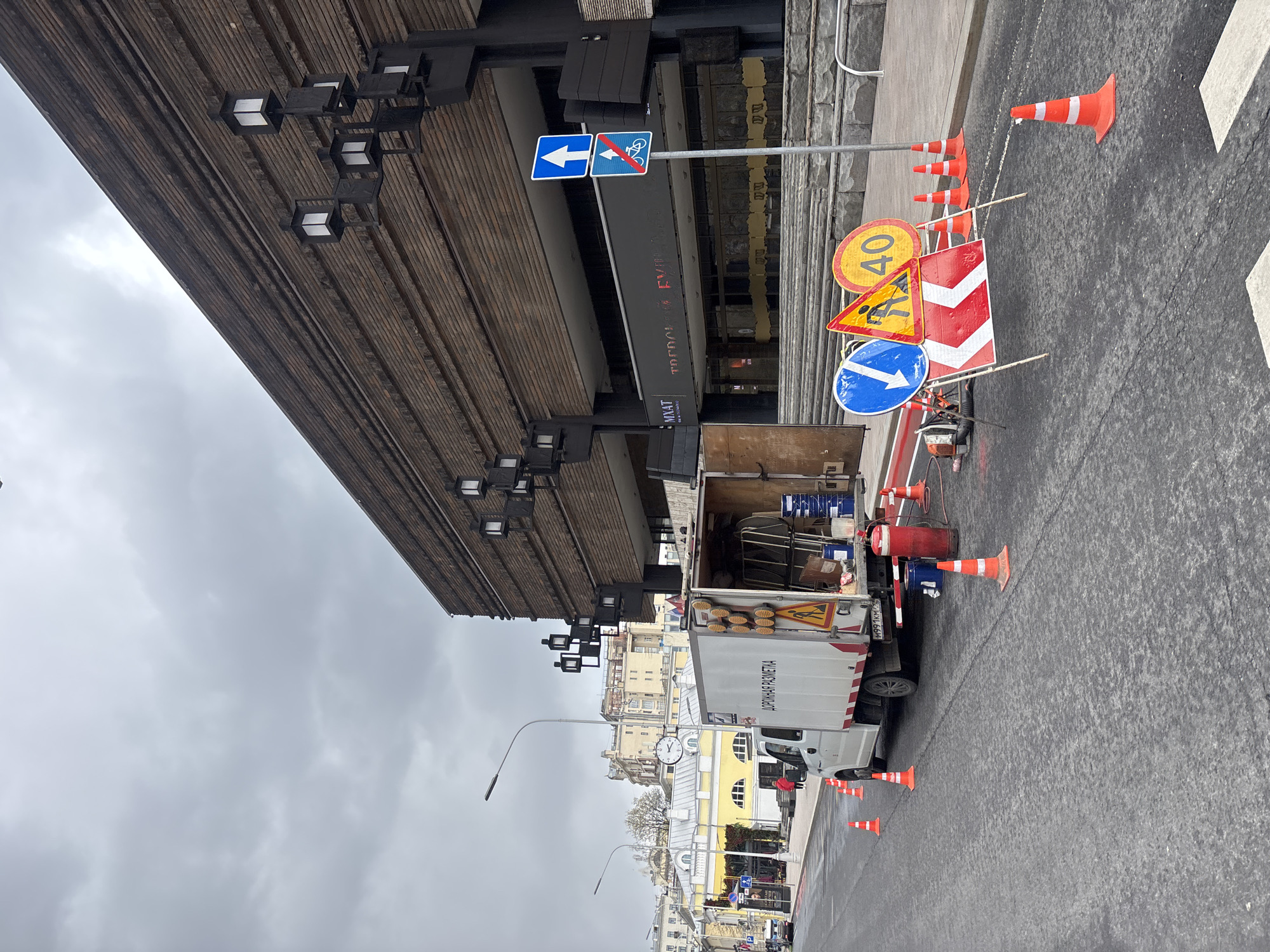 This image depicts a street scene in Moscow, Russia, indicating ongoing road work or maintenance under an overcast, grey sky.
In the foreground, a section of asphalt road is marked by numerous orange and white traffic cones. A white cargo truck is parked on the side of the road with its rear doors open, revealing various tools and equipment inside. The side of the truck clearly displays the Cyrillic text "ДОРОЖНАЯ РАЗМЕТКА," which translates to "ROAD MARKING," identifying its purpose.
A cluster of road signs is prominently displayed near the truck. These include a blue square sign with a white arrow indicating a left turn, a blue square sign prohibiting bicycles, a yellow circular speed limit sign showing "40," a yellow triangular warning sign depicting a person shoveling (signifying road work), a red and white chevron sign possibly indicating a hazard or diversion, and a blue circular sign with a white arrow pointing down-right, typically meaning "keep right." Adjacent to the truck, a red fire extinguisher, a blue barrel, and other road work machinery are visible.
To the left, a large building with a distinctive horizontally textured facade extends over the street. Its underside is fitted with modern, square-shaped light fixtures. In the background, a cityscape unfolds with other buildings, including a notable yellow structure with arched windows, and a public clock on one of the buildings. A person in a red jacket is faintly seen near the truck, and another individual appears to be working among the signs and equipment. The road surface appears damp, suggesting recent rain or wet conditions. The scene captures a moment of urban infrastructure upkeep.