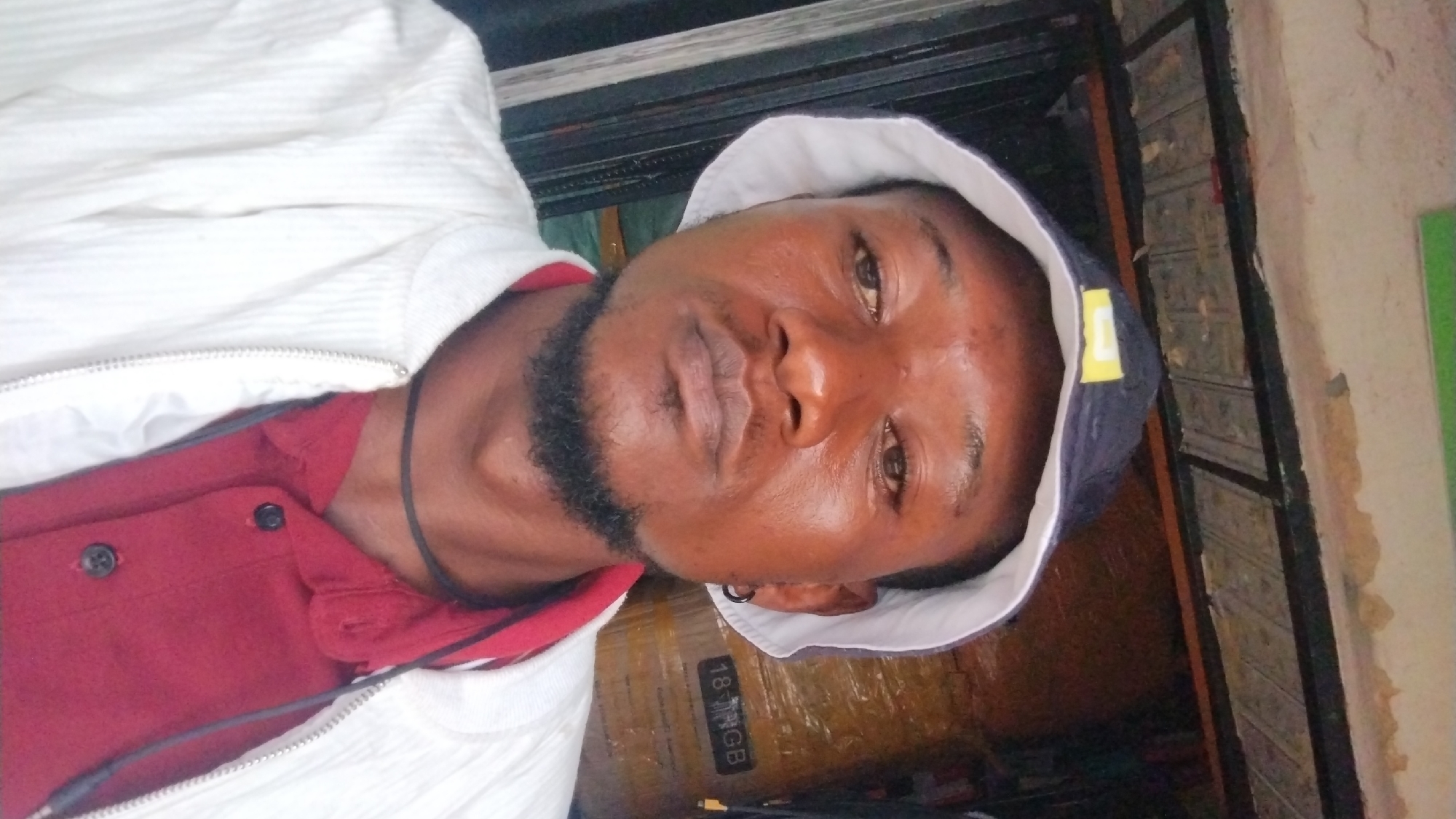 A man is centered in the frame, wearing a white jacket over a red button-down shirt and a dark bucket hat with a white lining. He has dark skin, a short beard, and an earring in his left ear. The background is partially obscured, but behind the man, there's a wrapped package with visible text "18 VGB", and structural elements like a metal frame or window bars. A wall with peeling paint and a green surface is seen to the right. The location is likely within the city of Jos, Nigeria.