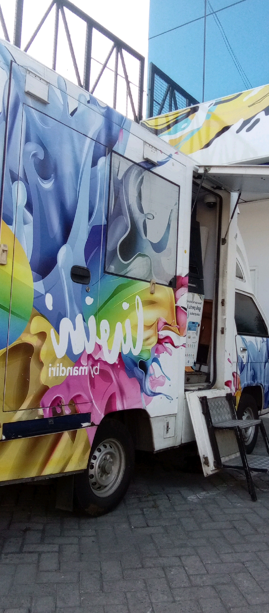 The image shows a white food truck with a colorful, abstract mural on its side. The mural features swirling shapes in shades of blue, yellow, pink, and green, with white text that appears to say "mvenil" and "inibnsmy" (likely a mirrored or stylized version of text). A door handle is visible on the side of the truck. The truck is parked on a paved surface, possibly a street or parking lot, in what appears to be an urban environment. In the background, there is a building with blue glass windows and a black metal structure. The scene is well-lit, suggesting it is daytime. The image does not contain any people.