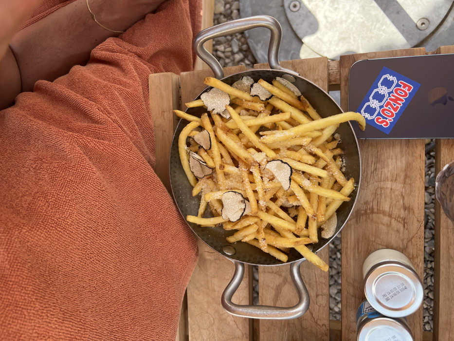 A metal pan filled with French fries topped with truffle shavings sits on a wooden surface next to an orange fabric item and a smartphone with a sticker that reads "FONZO'S." Two small cans are also visible on the wooden surface, and the surface appears to be made of parallel planks. The fries are golden and have a sprinkling of what appears to be salt or Parmesan cheese.