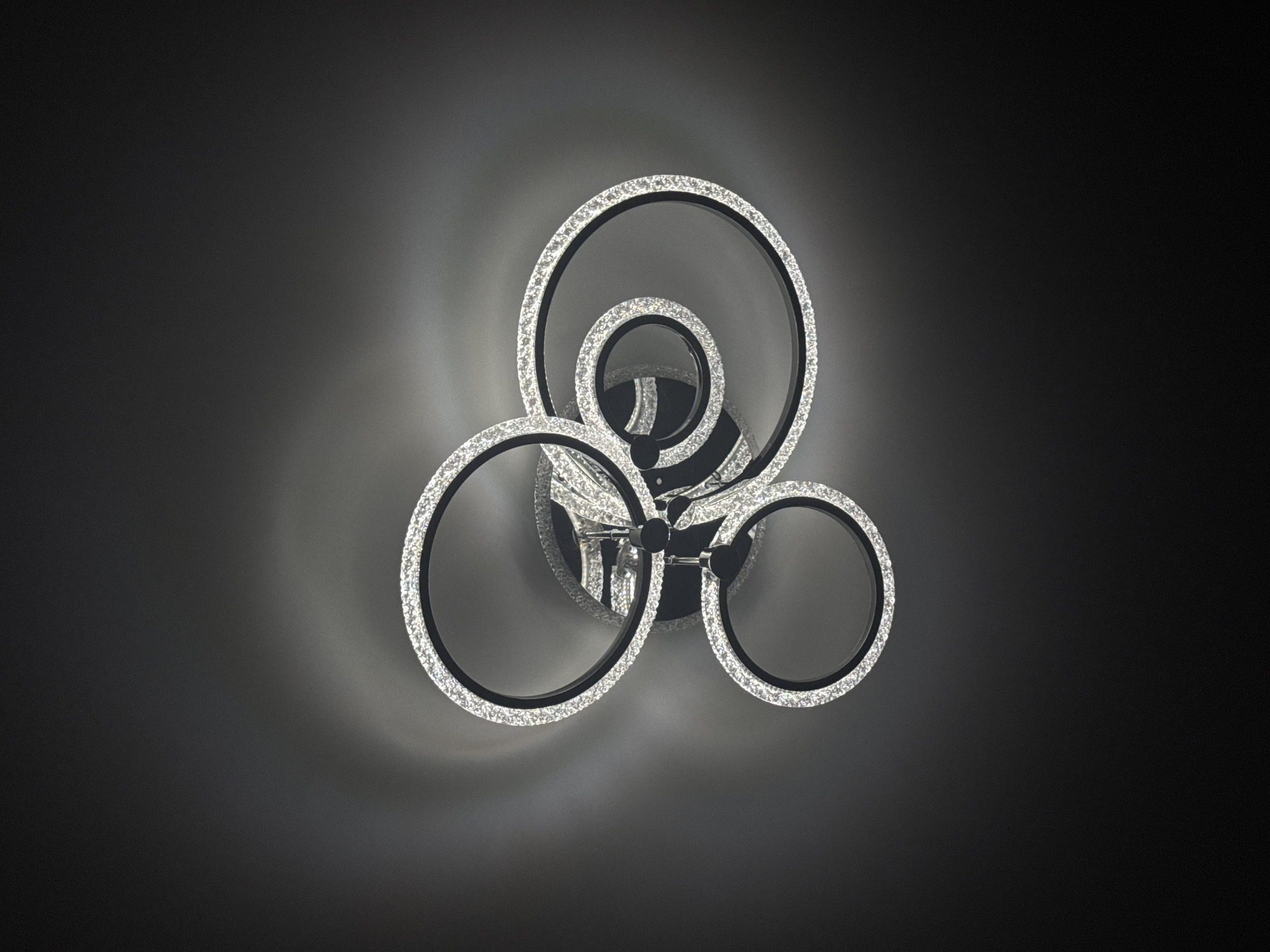 A ceiling-mounted light fixture is presented against a dark, smooth background. The fixture comprises multiple interconnected circular elements. Each ring features a dark, likely black, exterior frame and an interior surface densely adorned with small, highly reflective, faceted crystal-like components. These components emit a bright, diffuse white light, creating a luminous outline around each ring. The rings converge at a central, dark metallic mounting point. The light cast by the fixture illuminates the immediate surrounding ceiling area with a soft, circular glow that gradually diminishes into the deep grey and black tones of the background.