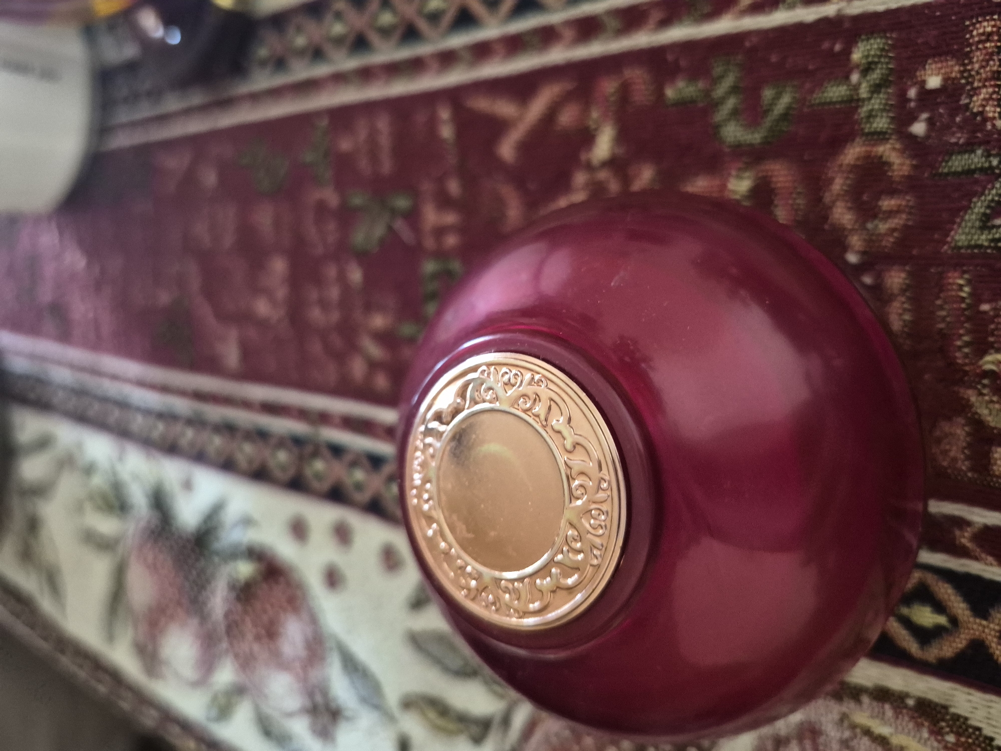 A close-up, high-angle shot captures a decorative, spherical object, possibly a perfume bottle or a small ornamental box, resting on a patterned fabric surface. The object itself is a vibrant reddish-purple, exhibiting a smooth, glossy finish that reflects ambient light. Adorning its top is a circular, gold-colored medallion with an intricately sculpted border featuring elaborate scrollwork and floral motifs. The center of the medallion is a smooth, reflective gold disk.
The object is positioned on a fabric, likely a tablecloth or runner, which displays rich, traditional patterns. The fabric features a broad central band of deep red with subtle, abstract designs. This central section is bordered by thinner strips showcasing geometric patterns, such as interlocking diamonds or zigzags, in muted tones of beige, brown, and hints of green and blue. To the left, a wider section of the fabric emerges with a lighter, off-white background, adorned with stylized illustrations of what appear to be pomegranates in reddish hues with green stems and leaves.
The image has a shallow depth of field, with the decorative object in sharp focus while the background fabric is slightly blurred. The lighting suggests an indoor setting. There are no people visible, and no specific activity is taking place; the scene is a static display of decorative items. No discernible text is visible on any part of the image. The traditional fabric patterns, particularly the prominent pomegranate motifs, are culturally significant and align with the provided location context of Yerevan, Armenia.