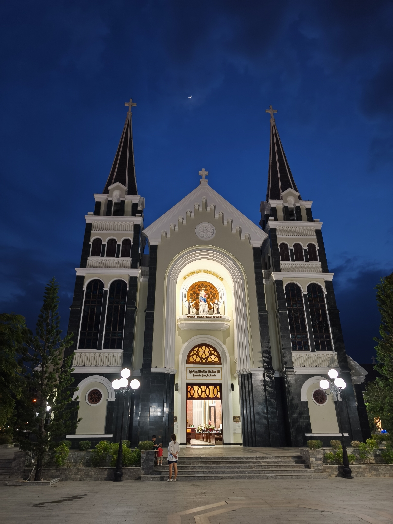 This is a night-time, low-angle, full-shot photograph of the Nha Trang Cathedral, also known as the Stone Church, located in the Bac Nha Trang Ward, Vietnam. The sky is a deep, dusky blue with a sliver of crescent moon visible near the top center. The cathedral is illuminated, highlighting its Gothic Revival architecture. It features two tall spires topped with crosses, a central facade with arched windows and a large rose window above the entrance. The entrance itself is brightly lit, revealing the interior. At the center of the facade, within an arched niche, is a statue of the Virgin Mary with the child Jesus. Two figures, a woman and two children, are standing on the wide, stone steps leading up to the cathedral entrance. The woman, wearing a white top and dark shorts, faces away from the camera. The two children, one in a red shirt and the other in a dark shirt, are also on the steps. The area in front of the cathedral is paved, and several lampposts with multiple globes cast pools of light on the ground. Lush green trees flank the sides of the cathedral, and small, manicured bushes line the lower parts of the building. The overall impression is one of serene grandeur at dusk.