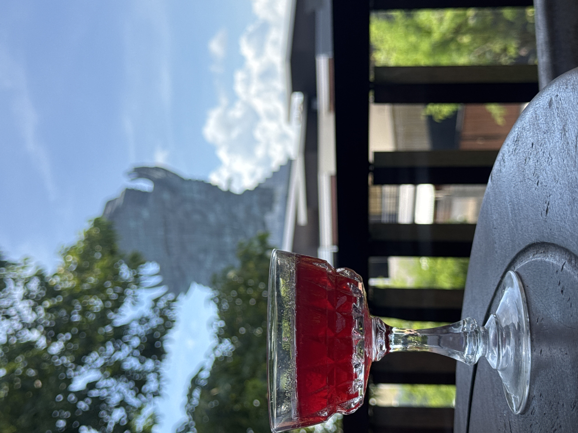 A tilted, eye-level shot features a glass of red liquid on a gray, round table. Outside the balcony, which has wooden railings, are lush, green trees, a building with gray siding, and a blue sky with white clouds. The scene is located in Ungasan, Indonesia.