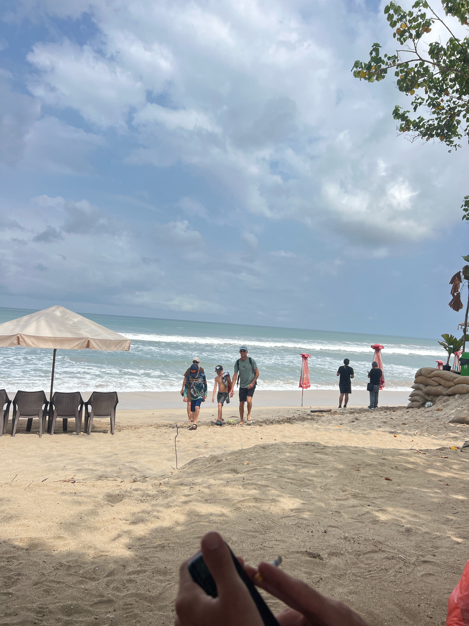 In Kuta, Indonesia, a beach scene unfolds under a cloudy sky. A large, light-colored umbrella shades a row of four empty, dark plastic chairs on the sand. Beyond the chairs, the ocean rolls with white-capped waves. Several people are walking along the wet sand near the water's edge. To the right, two red-topped umbrellas stand vertically, and a group of sandbags are piled up. A tree with green and yellowing leaves overhangs the scene from the upper right. In the foreground, a person's hand is visible, holding a lit cigarette.