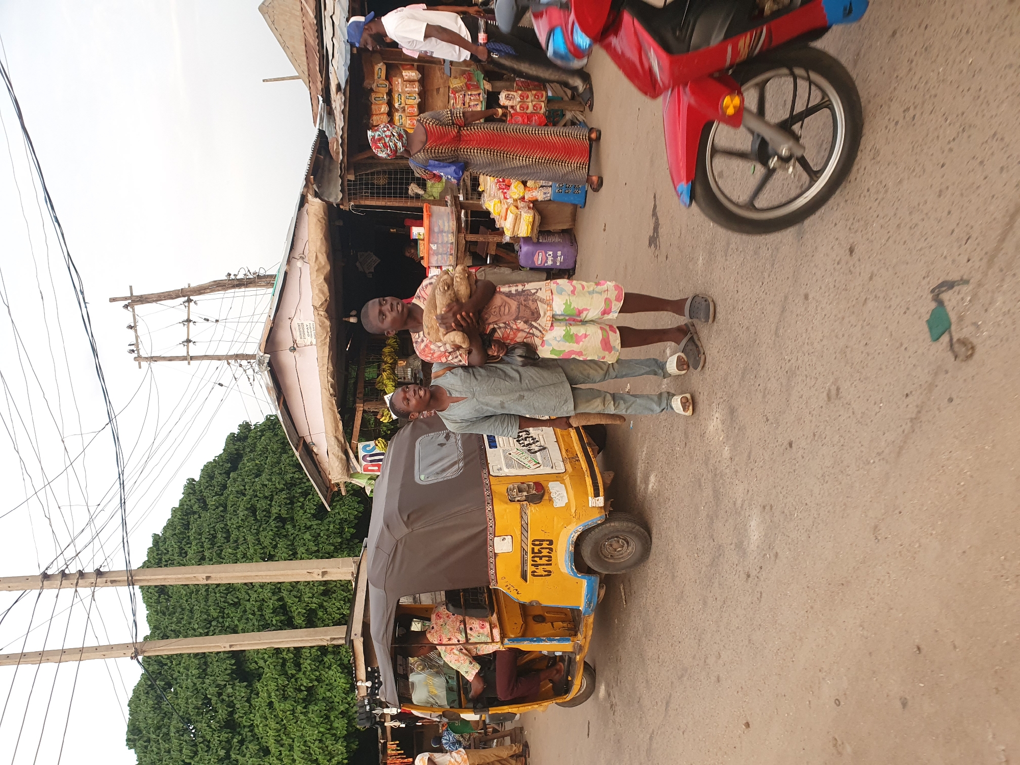The image depicts a street scene in Lokoja, Nigeria. Two boys stand in front of a yellow tricycle (keke) with a covered canopy. The tricycle has the number "C.1359" displayed on its side. The boy on the left wears a patterned shirt and shorts and holds what appears to be a bundle of items, possibly food. The boy on the right wears a blue shirt and pants and rests his arm on the tricycle. An older woman stands near a makeshift shop selling various goods, including bread and snacks. A man stands behind the shop near a red motorcycle. Power lines and poles are visible in the background, along with a lush green tree. The street is paved.