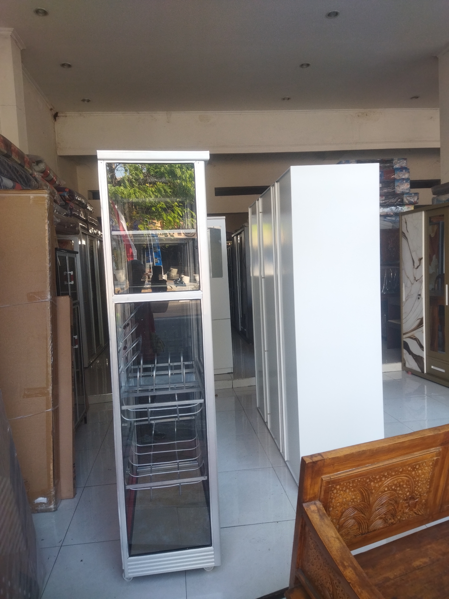 In Sukoharjo, Indonesia, a row of display cabinets and a white, rectangular cabinet are arranged in a retail space. The cabinets are made of aluminum and glass, with some having metal shelves. To the left of the cabinets is a tall cardboard box. A carved wooden bench is visible in the foreground. The environment suggests a furniture or appliance store.
