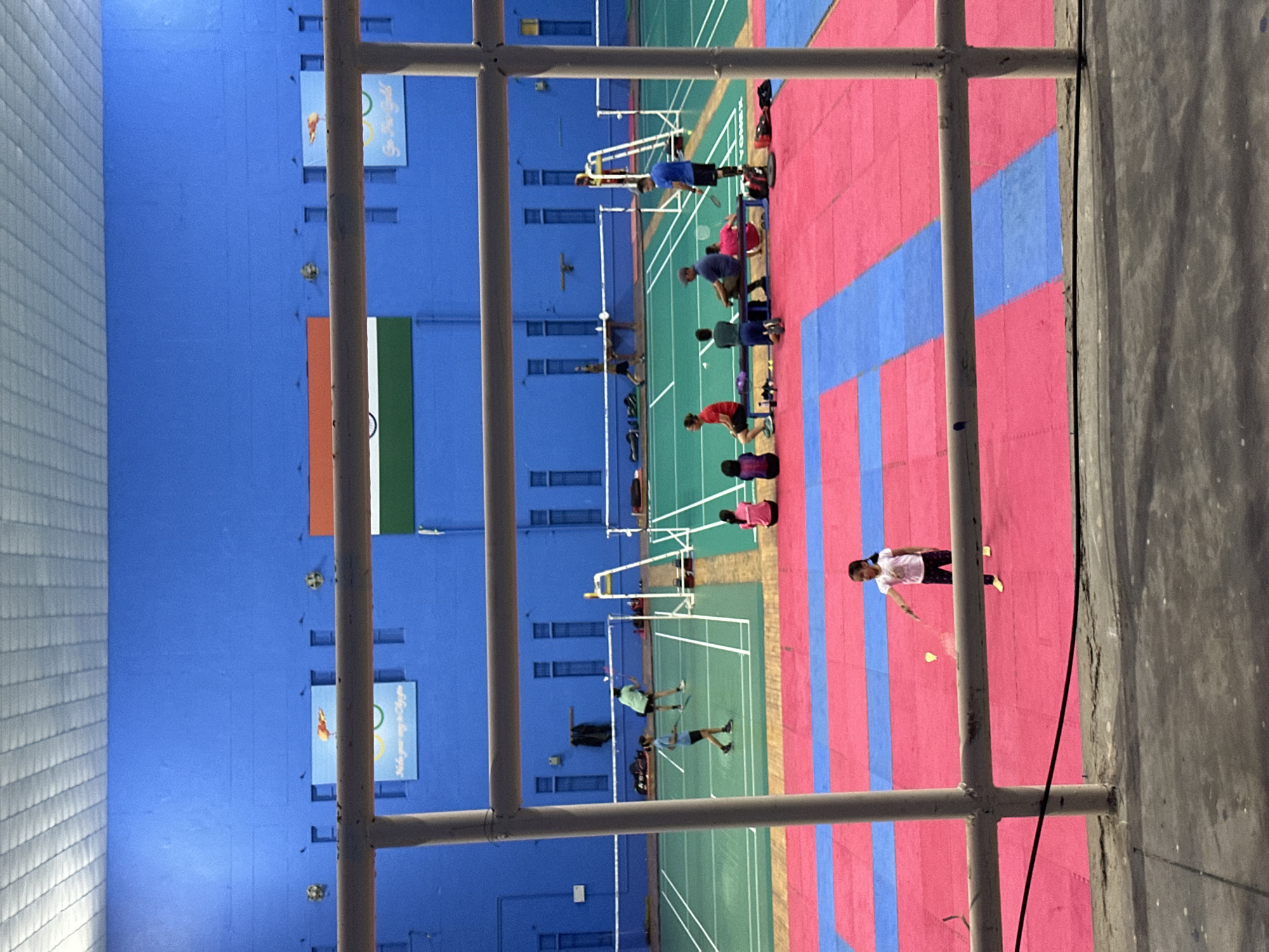 This image depicts the interior of an indoor badminton court. The court is divided into multiple playing areas marked with white lines on a green surface. The walls of the court are painted a bright blue. On the walls, there are several banners, including the Indian flag and a banner with Olympic rings.
In the foreground, there is a large metal railing system that partially obscures the view of the court. Beyond the railing, on the court, several people are visible. On one of the courts, two young girls are playing badminton, one is serving. Further back, on another court, a group of people are sitting on a bench, observing the game. Some individuals are actively playing badminton, and others are spectators.
The floor of the court is covered with brightly colored mats, predominantly pink and blue, in a checkered pattern. A young girl in a white shirt and dark pants is standing on the pink mat, holding a badminton racket and about to hit a shuttlecock. The lighting appears to be artificial, coming from overhead fixtures. The overall impression is of a busy sports facility.