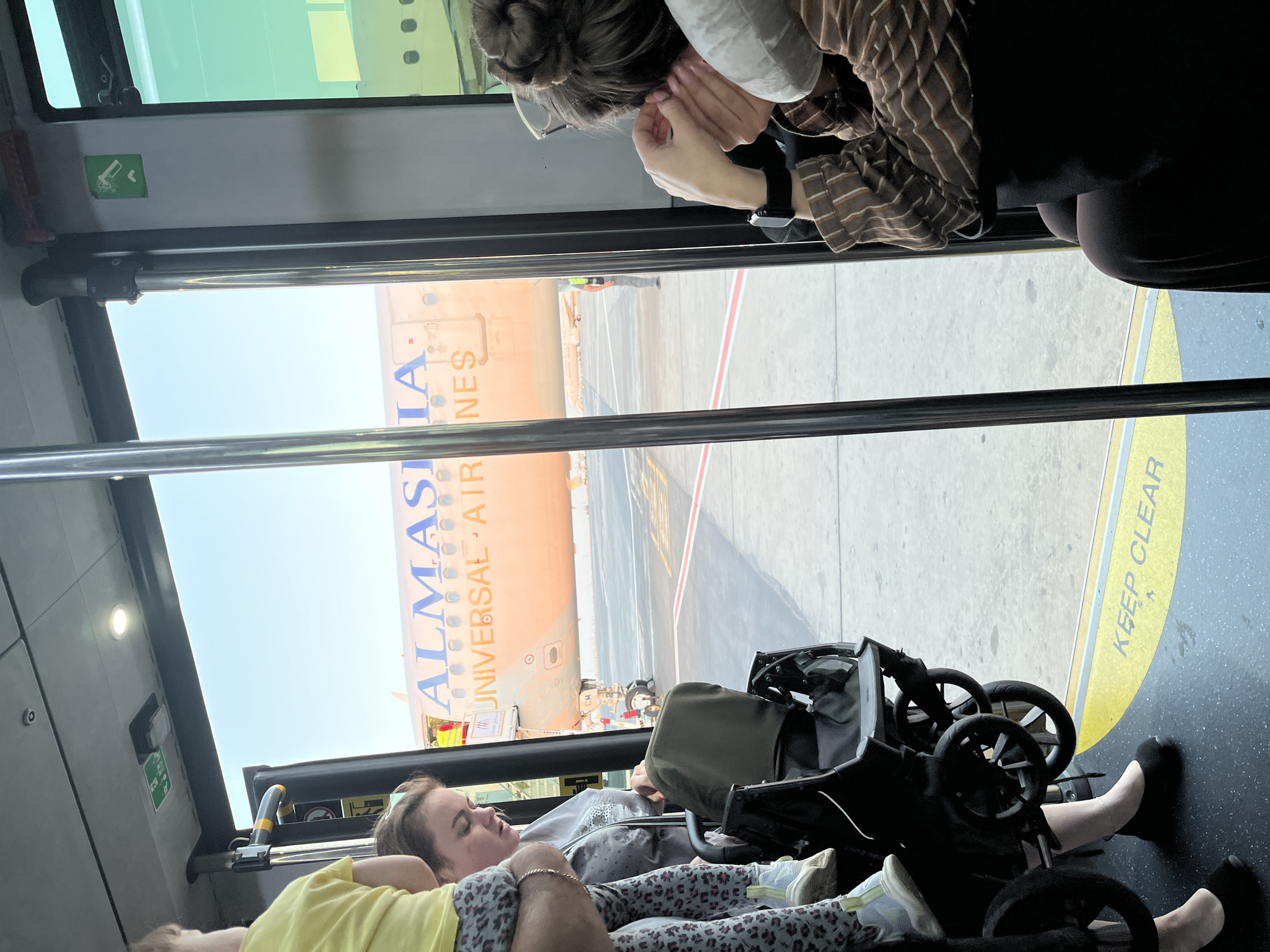 The image shows a view from inside a bus, looking out towards an airplane parked on the tarmac at an airport. The bus appears to be a passenger transport vehicle, possibly for boarding or disembarking an aircraft. Inside the bus, several people are visible, including a child in a yellow shirt, a woman with her hair pulled back, and another woman in a striped shirt. A stroller is also present in the bus. On the side of the airplane, the text "ALMASIA" and "UNIVERSAL AIRLINES" can be clearly read. The ground outside the bus is a paved surface, likely an airport apron, with yellow lines and markings visible. The sky is clear and blue. The scene suggests passengers are being transported between the terminal and the aircraft.