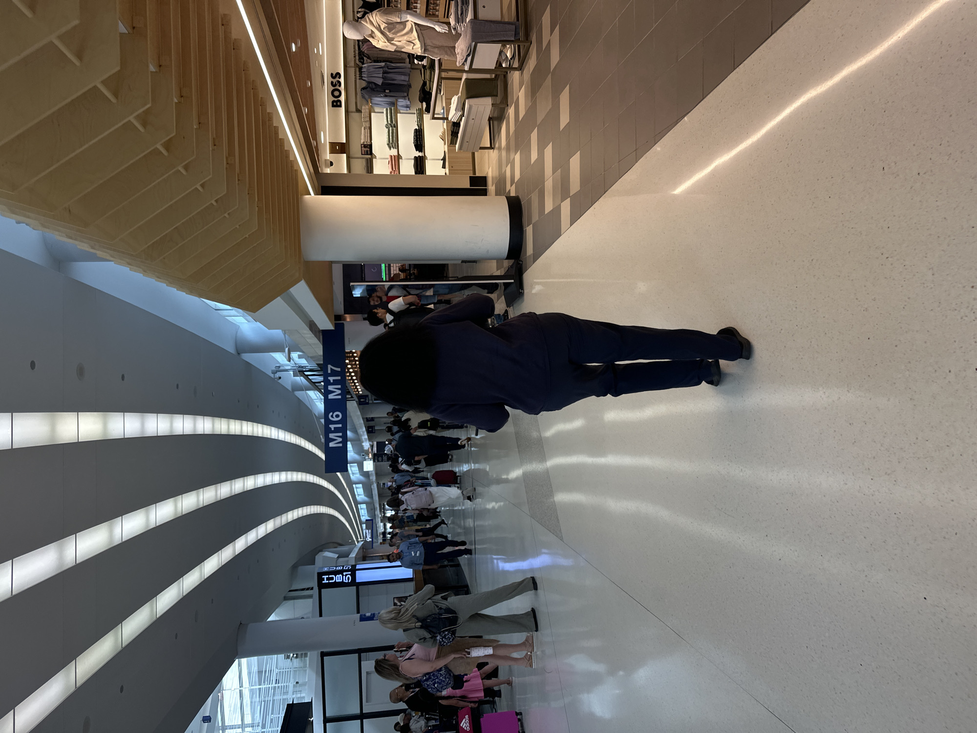 The image captures a busy airport concourse, likely at O'Hare International Airport in Chicago, Illinois. The architecture features a modern design with a curved ceiling illuminated by long, recessed light fixtures. In the foreground, a person with dark, curly hair is walking away from the camera, wearing dark pants and a dark top.
Further down the concourse, several other individuals are visible, some walking, some standing. To the left, a group of three women are standing, one in a pink dress and two in casual attire with backpacks. Luggage, including pink suitcases, is scattered around the area.
Signage for gates M16 and M17 is prominently displayed overhead. On the right side of the concourse, a retail store with a "BOSS" logo is visible, showcasing men's clothing on display racks and mannequins. The floor is a polished white speckled material, transitioning to a darker tiled area further down the concourse. The overall atmosphere suggests a typical airport environment with travelers moving through the space.