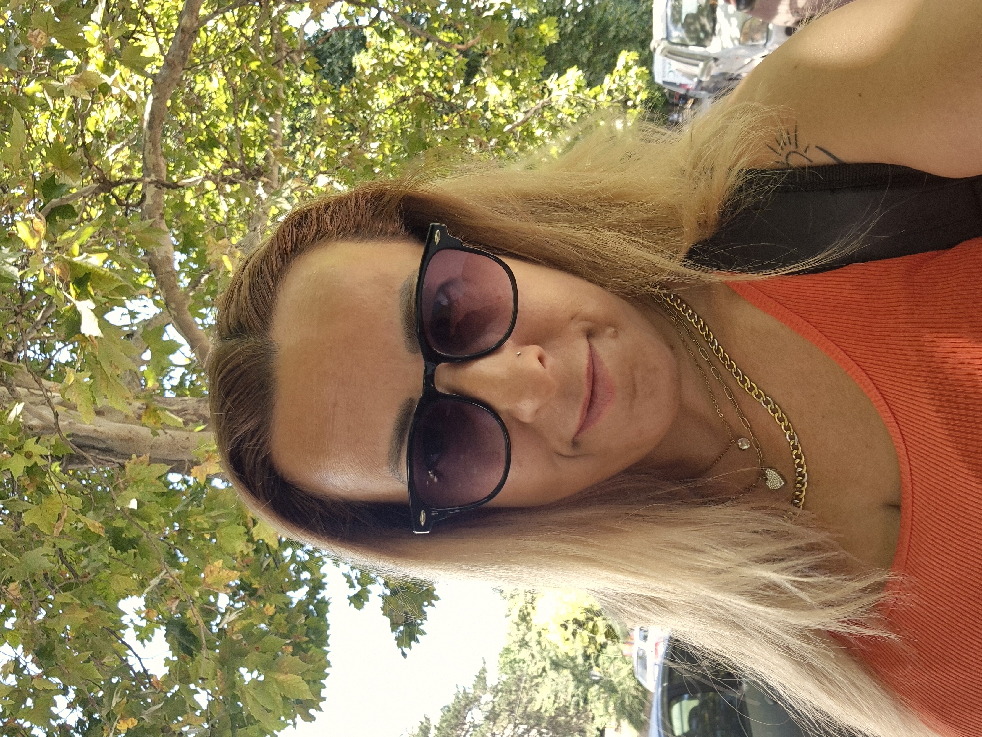 A medium shot taken from a slightly low angle shows a woman with blonde hair and sunglasses, smiling at the camera. She is wearing an orange tank top and multiple necklaces, one of which has a heart pendant. A tattoo is visible on her right arm. The background is a blurry mix of trees and some parked cars, suggesting an outdoor setting, possibly a park or a street. The lighting indicates it is daytime.