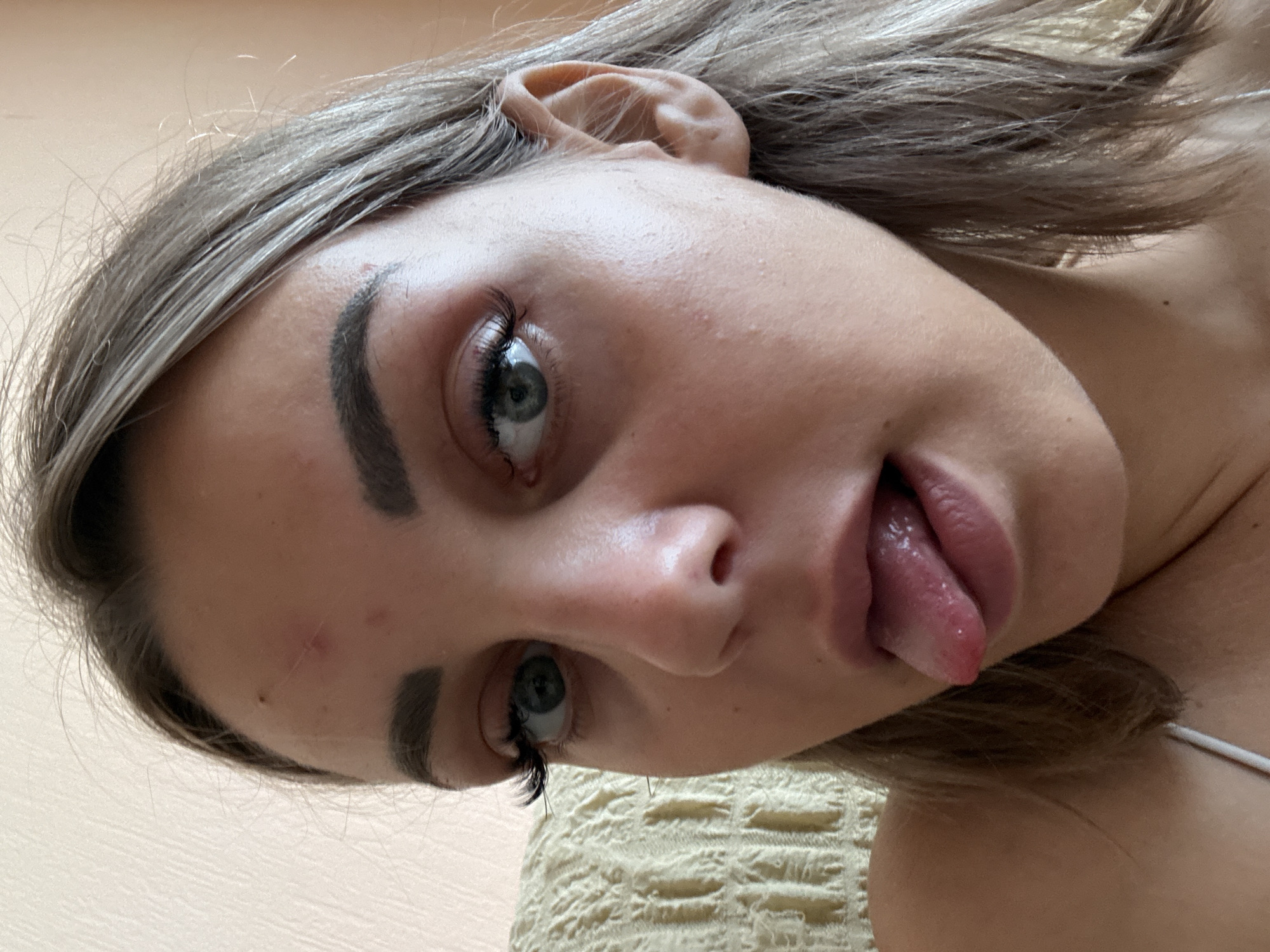 The image is a close-up, selfie-style portrait of a young woman with blonde hair. She is looking towards the camera with her tongue extended. Her face is the primary subject. She has dark eyebrows, prominent eyelashes, and light-colored eyes. There is a small scar or mark on her forehead. The background is a textured, light-colored wall and a portion of a light-colored, textured fabric that appears to be upholstery. The lighting suggests an indoor environment, possibly daytime. The image does not contain any information that would allow for the determination of its location in Kholmsk, Russia, or any specific city.