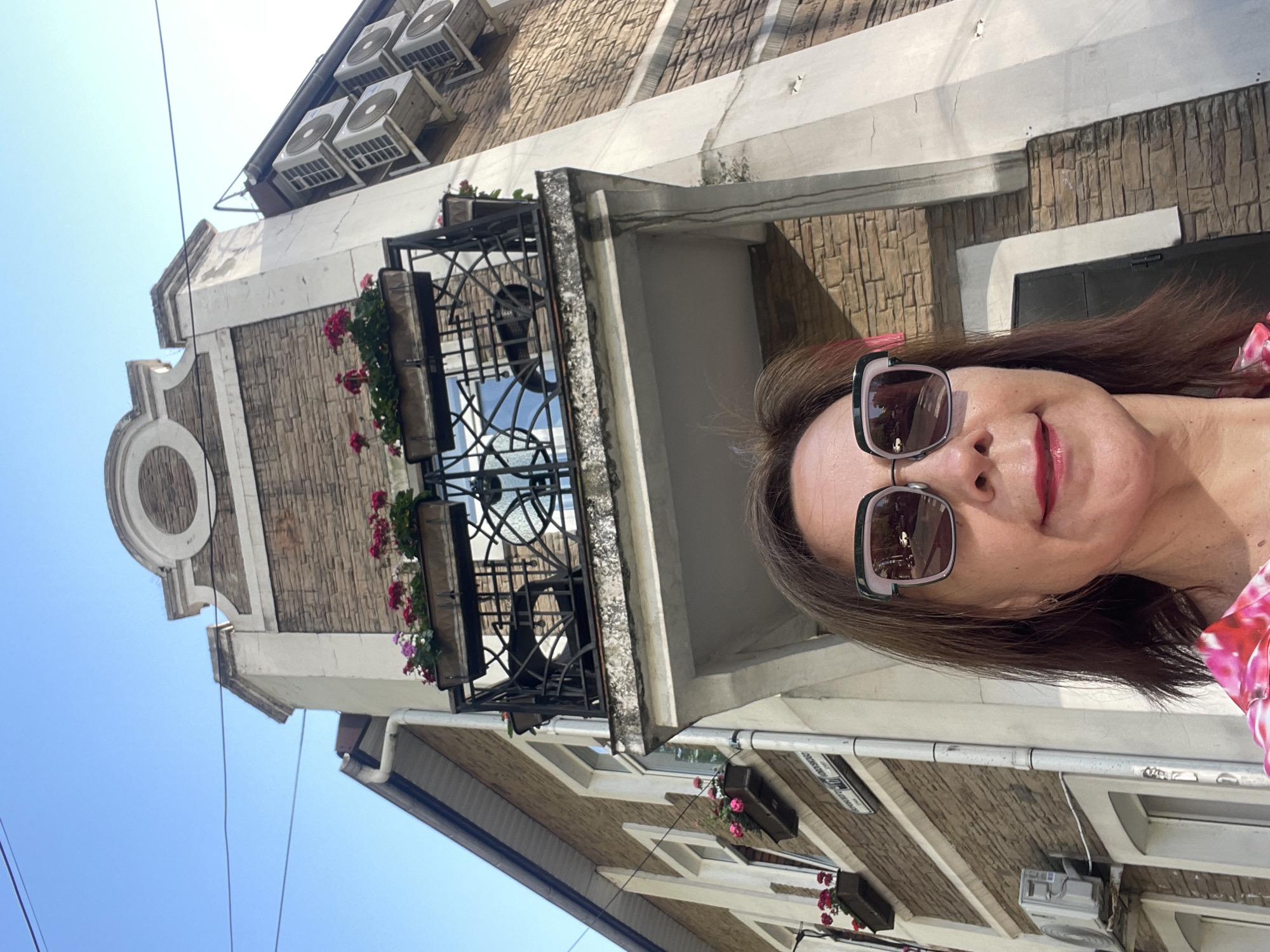 A medium shot captures a woman in the foreground, looking directly at the camera. She has brown hair and is wearing large, square sunglasses with dark lenses that reflect the street scene. Her lips are painted with a pink lipstick. She is wearing a bright pink and white patterned top. Behind her, the perspective shifts to an upward view of a building with a brick facade. Above the woman's head, a balcony is visible with railings and flower boxes filled with pink and white flowers. Further up, several air conditioning units are mounted on the side of the building. The sky above is clear and blue. The scene appears to be on a street in Sochi, Russia, indicated by a street sign visible on the building.