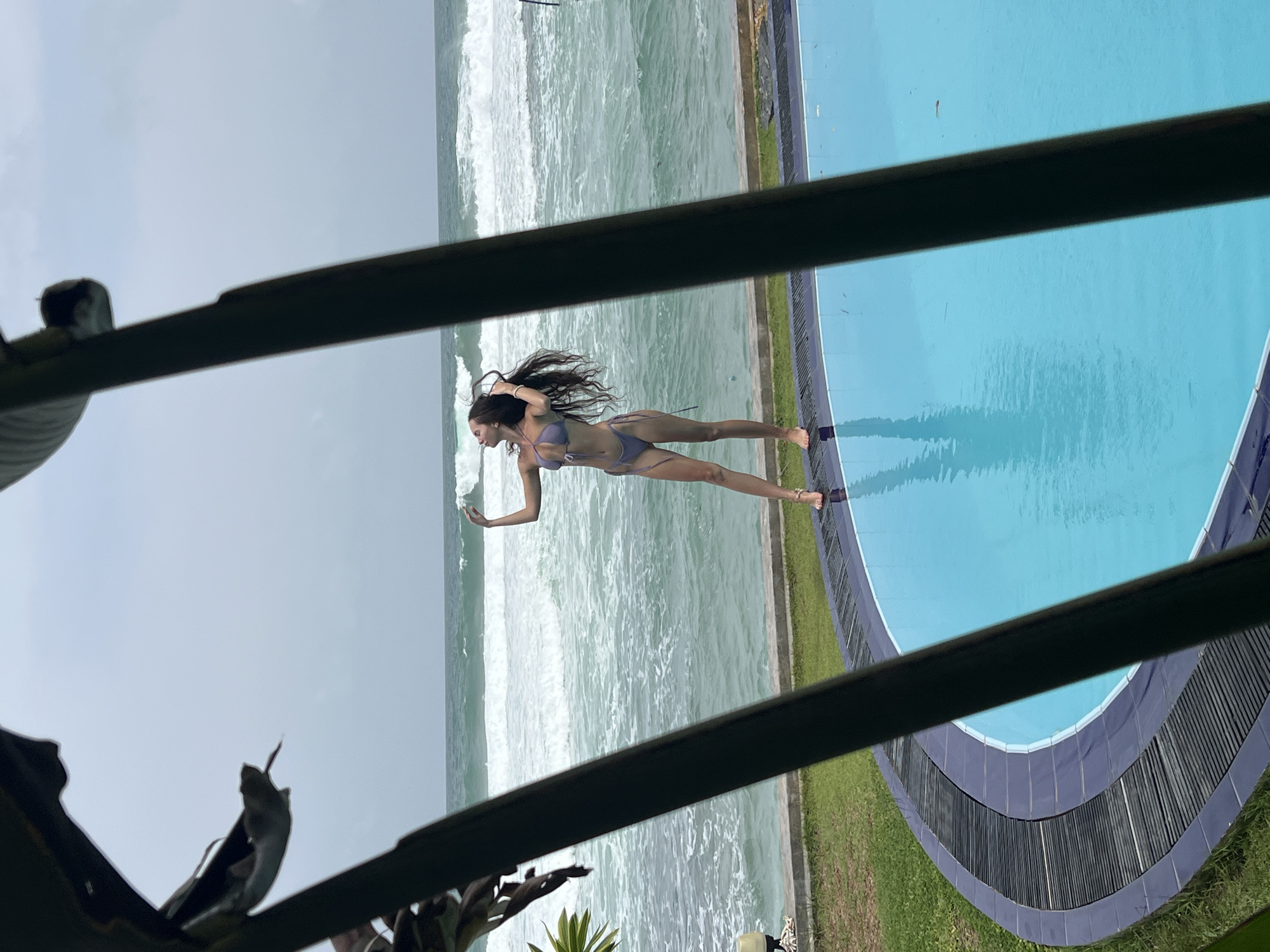 A horizontally oriented medium shot taken from an elevated perspective through foliage shows a woman in a lavender bikini standing between a pool and the ocean in Goviyapana, Sri Lanka. The woman is facing the ocean, with her left arm raised and extended, seemingly holding a device. The ocean is turbulent with whitecaps. A curved pool borders the ocean, with green grass separating it from the pool. Foliage is in the foreground. The sky is overcast.