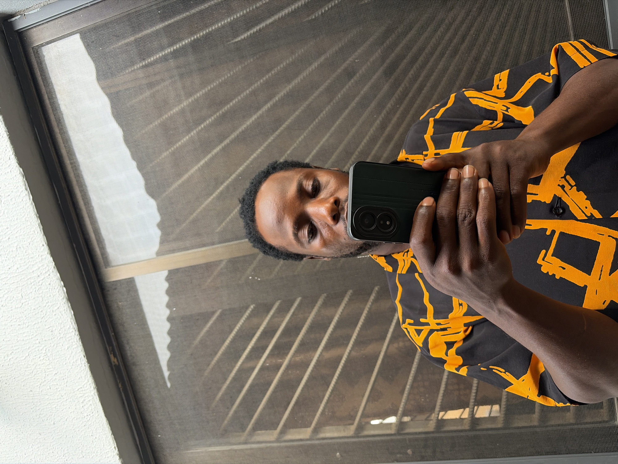 A man holds a smartphone with dual rear cameras in front of his face, seemingly taking a selfie. He is wearing a black shirt with an orange pattern. The background appears to be a window screen with a view of what might be a corrugated metal structure outside. The location is potentially Jos, Nigeria, inferred from the description request but not directly visible within the image. The image suggests an indoor setting with natural light coming through the window.