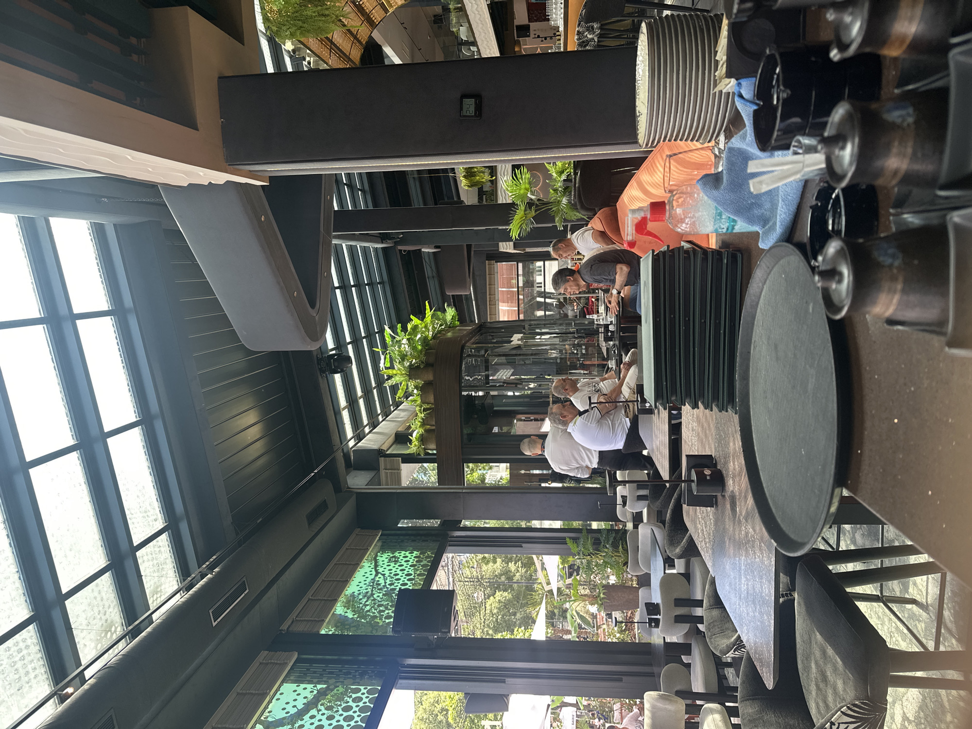 The image depicts the interior of a modern restaurant with an open-air feel due to a large glass roof and windows. Several patrons are seated at tables, engaged in conversations or dining. The decor is contemporary, with dark pillars, gray upholstery on chairs, and an abundance of potted plants. A counter area is visible in the foreground, featuring stacks of plates, a spray bottle, a blue cloth, and various containers. The overall atmosphere suggests a relaxed and upscale dining establishment. The specific location within the city is not identifiable from the image.