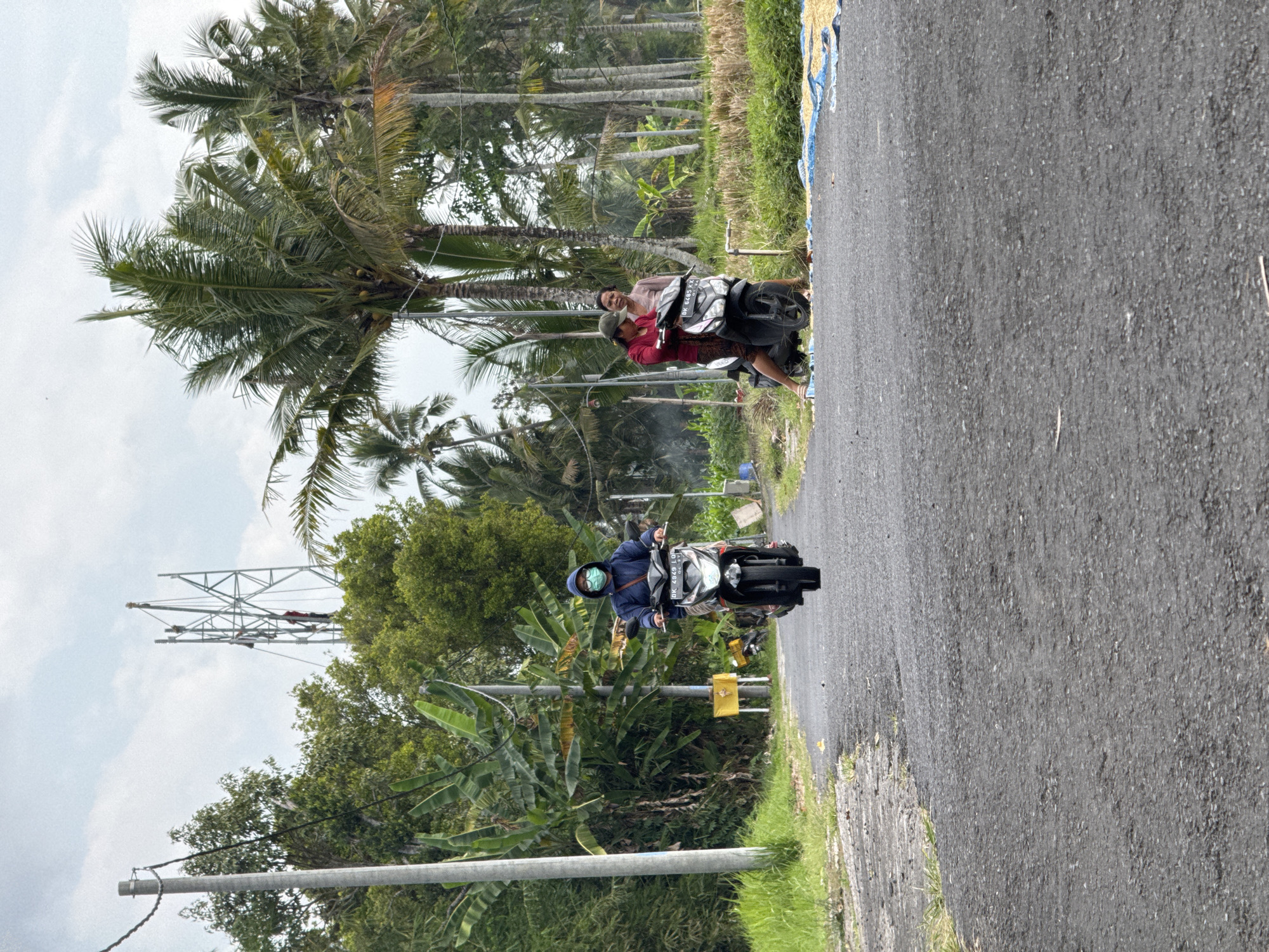 In Gianyar, Indonesia, on a rural, paved road lined with lush vegetation and tall palm trees, a person wearing a blue hoodie and a light blue face mask rides a black motorcycle towards the camera. They appear to be alone on the motorcycle. Further down the road, two people, one wearing a red shirt and the other a light brown shirt, are stopped on a black motorcycle. The person in the red shirt is looking away from the camera, while the person in the brown shirt is looking towards the camera. In the background, a large metal tower is visible, with several individuals working on its structure. The sky is overcast, with a few patches of blue visible. The environment suggests a tropical climate.