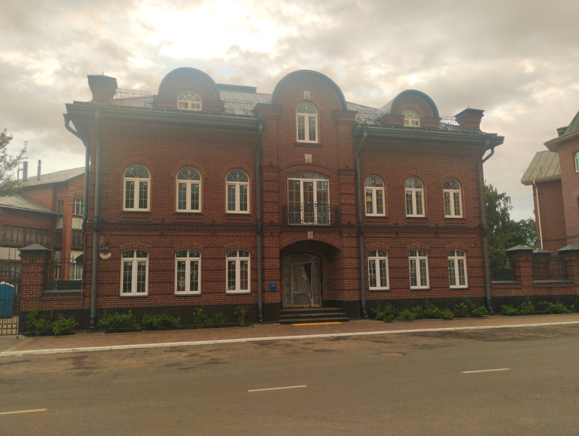 A two-story, red brick building with arched windows and a dark gray roof stands on a paved street. The building has a central arched entryway with steps leading up to it. A small blue porta-potty is visible behind a black metal fence on the left side of the building. The sky is cloudy and overcast. The building is located in the city of Kostroma, Russia.