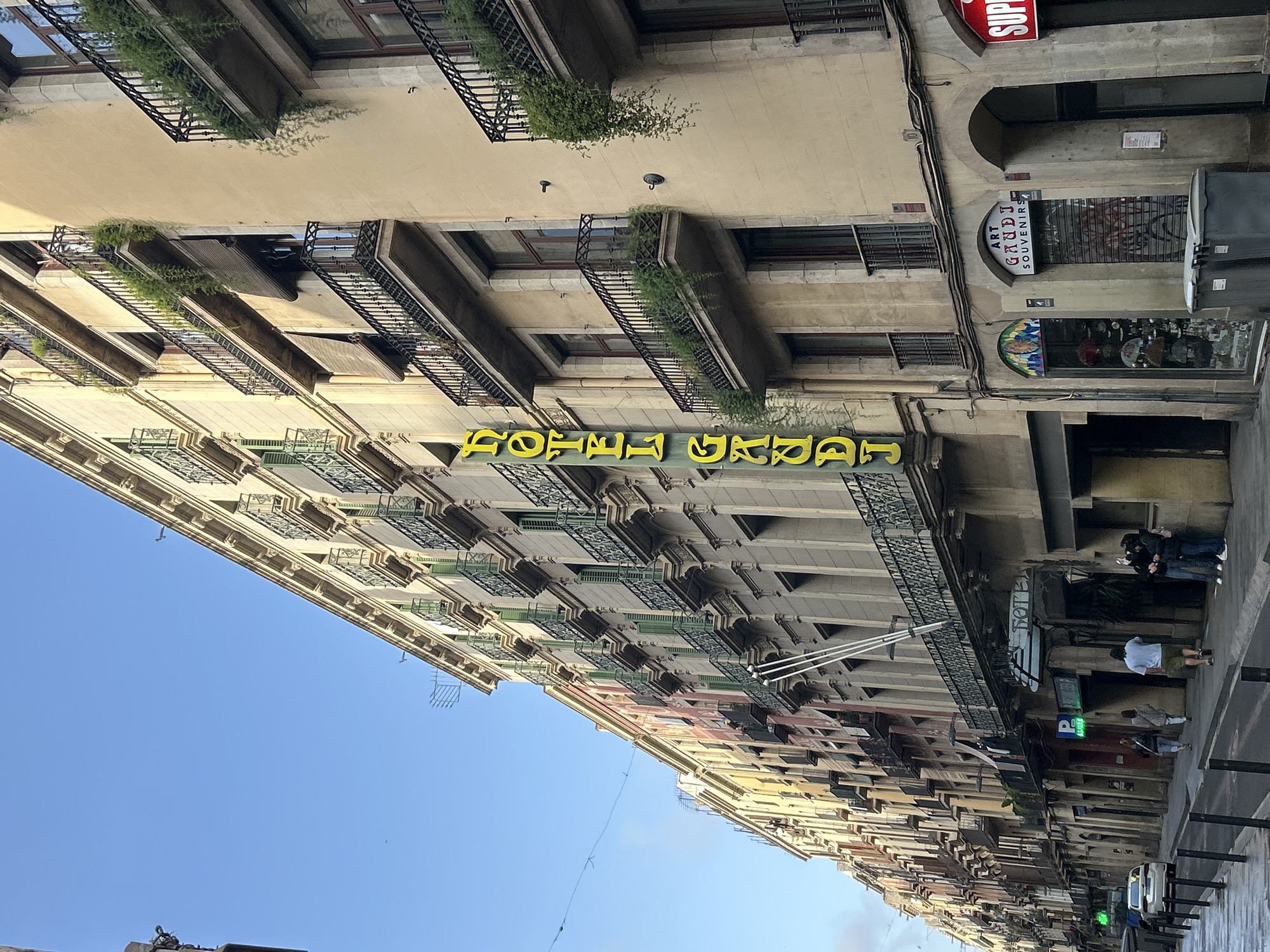 This photograph captures a vibrant street scene in Barcelona, Spain, looking upwards at a row of multi-story buildings under a clear blue sky. The buildings are characterized by their cream-colored facades and classic European architecture, featuring numerous windows and intricate wrought-iron balconies. Many of these balconies are adorned with lush green plants, adding a touch of nature to the urban environment.
A prominent yellow neon sign on one of the main buildings clearly reads "HOTEL GRAP," indicating a hotel establishment. At street level, below and to the right of this sign, a shop with a distinctive circular sign proclaims "ART GAUDI SOUVENIRS," a strong cultural indicator of Barcelona, given Antoni Gaudí's architectural legacy in the city. This souvenir shop features a display window with various items, and a partially visible red sign further down the street reads "SUP..." likely part of a "SUPER" market sign. Other street-level features include an arched doorway, metal grates on windows, a dark grey public waste bin, and a green sign displaying a white 'P' symbol for parking. Electrical wires are visible running along the building facades.
Several individuals are present on the sidewalk below, engaged in typical urban activities. Two people are standing near the "ART GAUDI SOUVENIRS" shop, and further along the street, a few more pedestrians are visible, some appearing to be interacting with their mobile phones.
The scene is illuminated by bright, clear daylight, suggesting either late morning or early afternoon under sunny weather conditions. The overall impression is one of a lively, characteristic European city street, with the "ART GAUDI SOUVENIRS" sign serving as a definitive location cue for Barcelona.