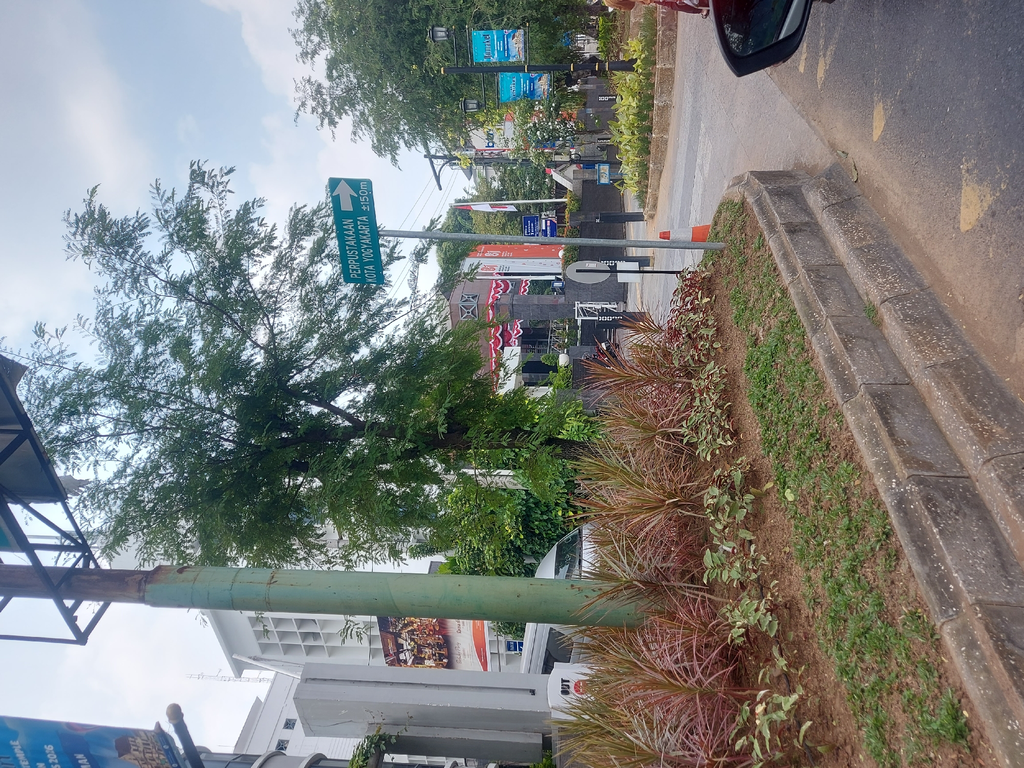 The image shows a street scene in Yogyakarta, Indonesia. In the foreground, there is a curb with a planter box containing various plants, including reddish-brown spiky plants and smaller green leafy plants. A tall, weathered metal pole is visible in the center left of the frame, with a green directional sign attached to it. The sign indicates "PERPUSTAKAAN KOTA YOGYAKARTA ±150m" with an arrow pointing to the right, suggesting the location of the Yogyakarta City Library. Behind the planter box and pole, there is a building with white and grey architectural features and some trees. Further in the background, more buildings, trees, streetlights, and a large flag can be seen. A person's legs and feet are visible on the right side of the image, suggesting they are standing near a car. The sky is partly cloudy.