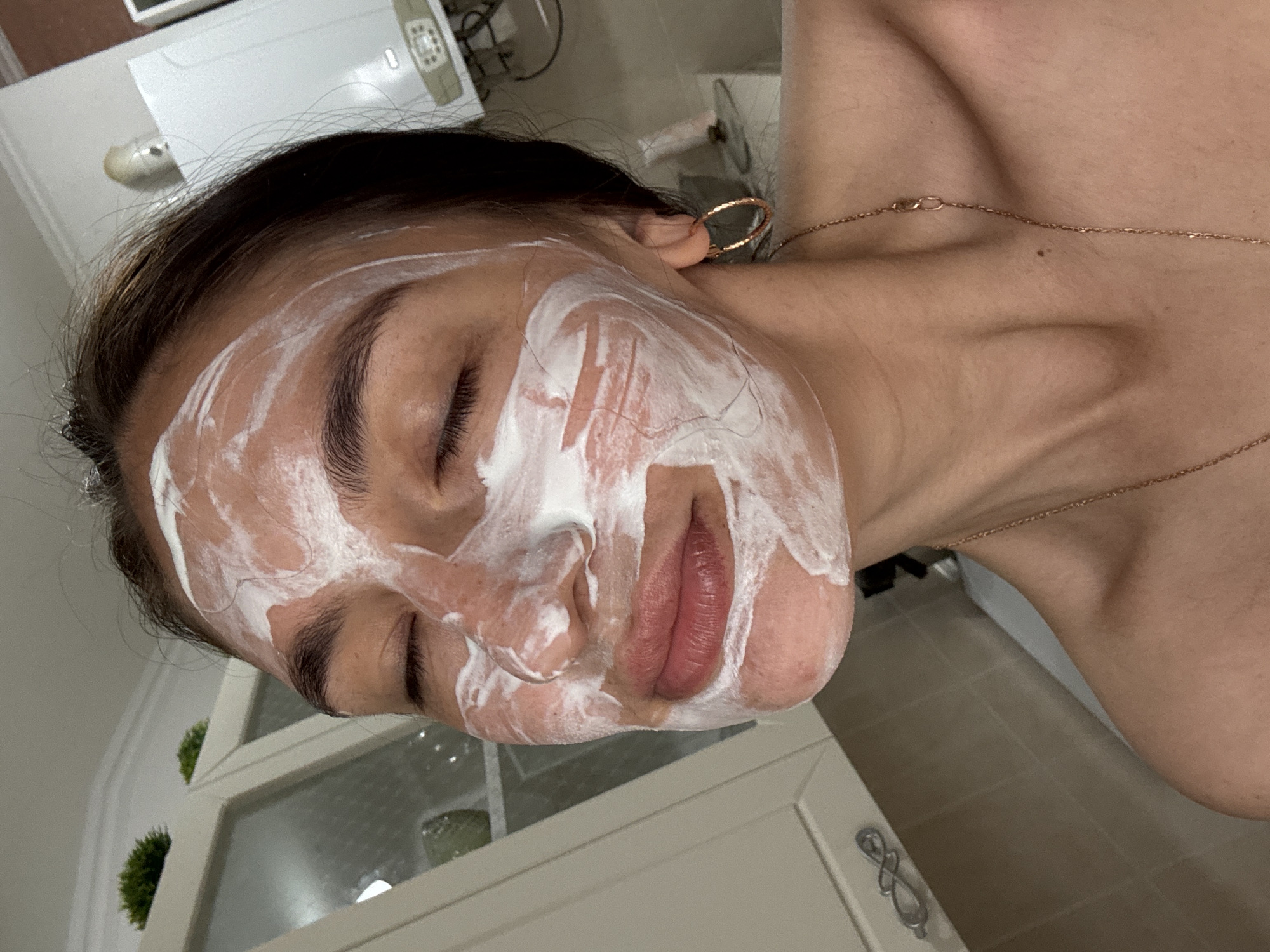A person with closed eyes has a white facial mask on their face. They are wearing a gold necklace and a gold hoop earring. The background includes a cabinet, a white appliance, and tiled flooring. The location in the city of Sochi, Russia is not discernible from the image.