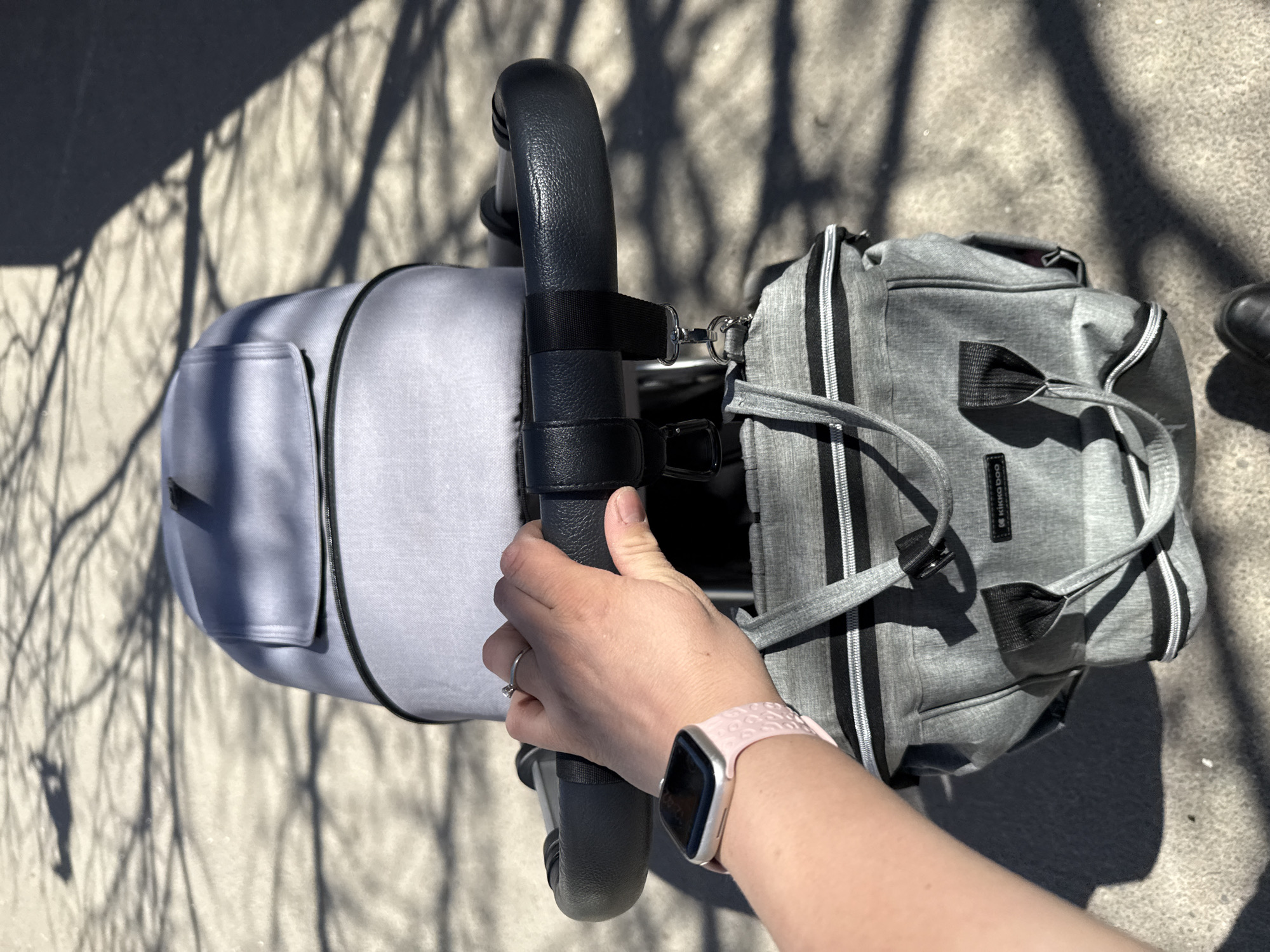 A person pushes a gray stroller with a gray diaper bag attached in an outdoor location. The stroller has a black handle and frame, and the diaper bag is attached to the handle with clips. The person's hand is holding the stroller handle, and they are wearing a pink watch. The ground is concrete and partially covered in shadows from a tree. The location could be Sofia, Bulgaria, but there is no specific evidence to confirm this.