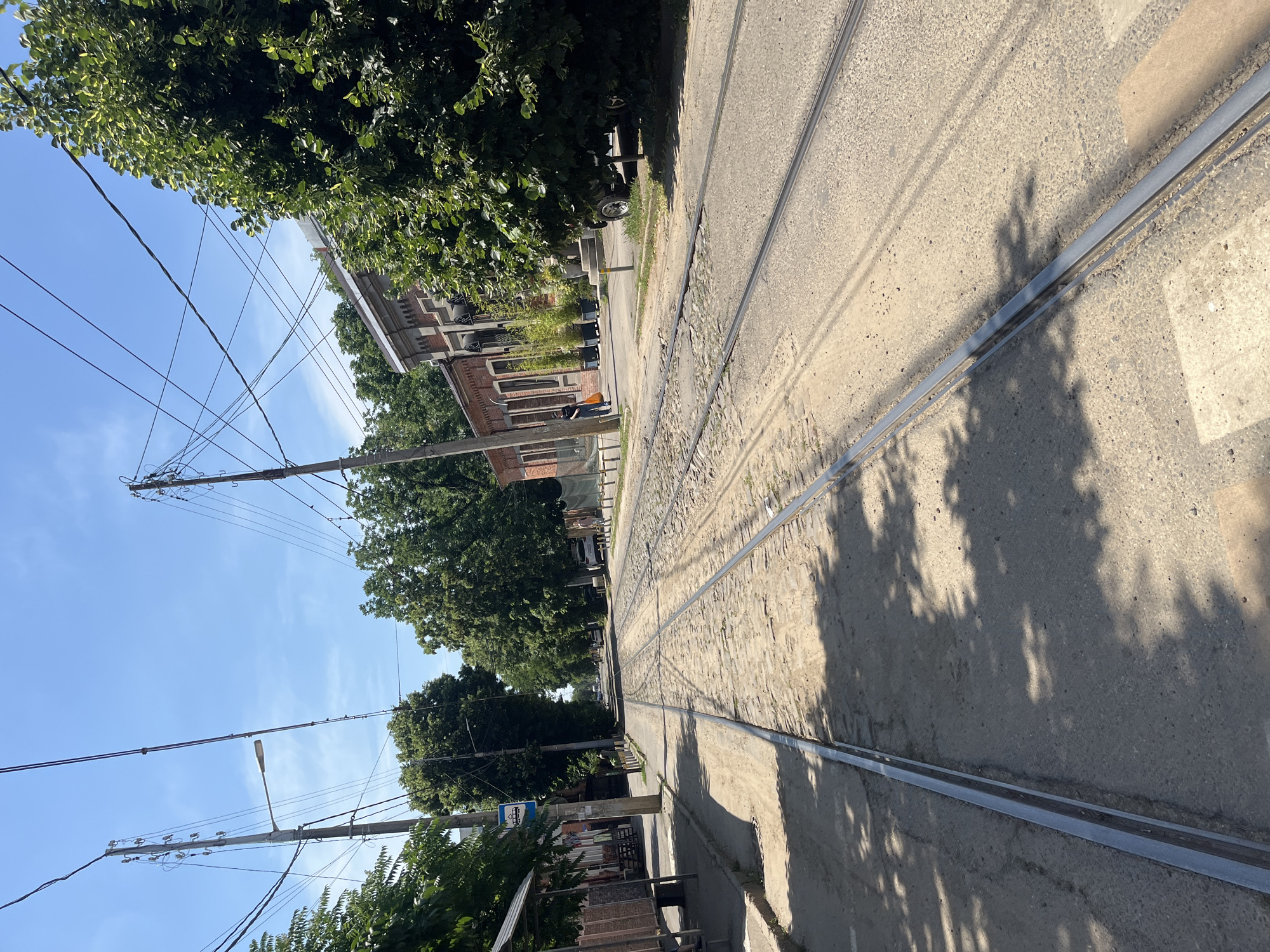 The image depicts a street scene in Krasnodar, Russia, characterized by tram tracks running down the center of the road. The tracks are laid on a cobblestone and asphalt surface, with some areas showing signs of wear and tear. Lush green trees line both sides of the street, casting dappled shadows on the ground.
On the left side of the street, a bus stop shelter with a blue and white sign is visible. Beyond the shelter, there are buildings with storefronts. Utility poles with a complex network of wires are a prominent feature, indicating overhead power lines for streetcars or electricity.
On the right side of the street, a brick building with arched windows stands. In front of this building, there are potted plants and a dark-colored vehicle, possibly a Jeep, parked under the shade of a tree. A person in a black shirt and light-colored pants is walking away from the viewer towards the brick building, carrying an orange bag. Further down the street, another person is visible, walking alongside a white car parked on the side of the road.
The sky is a clear blue with a few wispy clouds, suggesting a sunny day. The overall impression is of a somewhat old-fashioned urban environment, likely a residential or mixed-use area, where the tram system is still an active mode of transportation.
