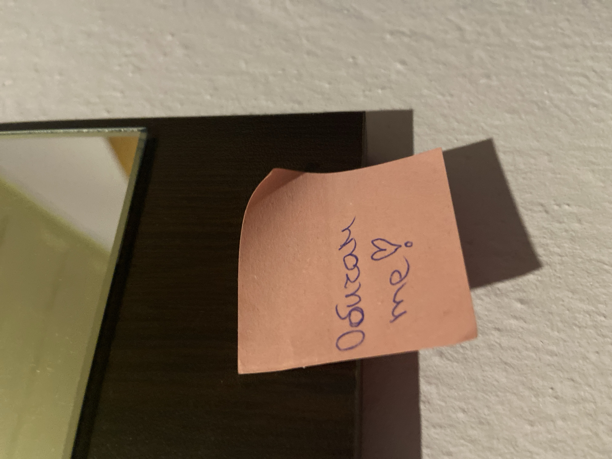 A close-up, slightly angled shot captures a pink sticky note adhered to a textured, off-white wall. The sticky note has purple handwriting in Cyrillic script that reads "Обичам те ♡" which translates to "I love you ♡". The note is positioned in the lower left quadrant of the frame, casting a shadow to its right. In the upper portion of the image, a dark, reflective surface, likely a mirror or a dark framed object, is visible, taking up the top half of the frame. The background environment suggests an indoor setting, possibly a residential or office space. There is no information or visual clue indicating the specific location in the city of Mindia, Bulgaria.