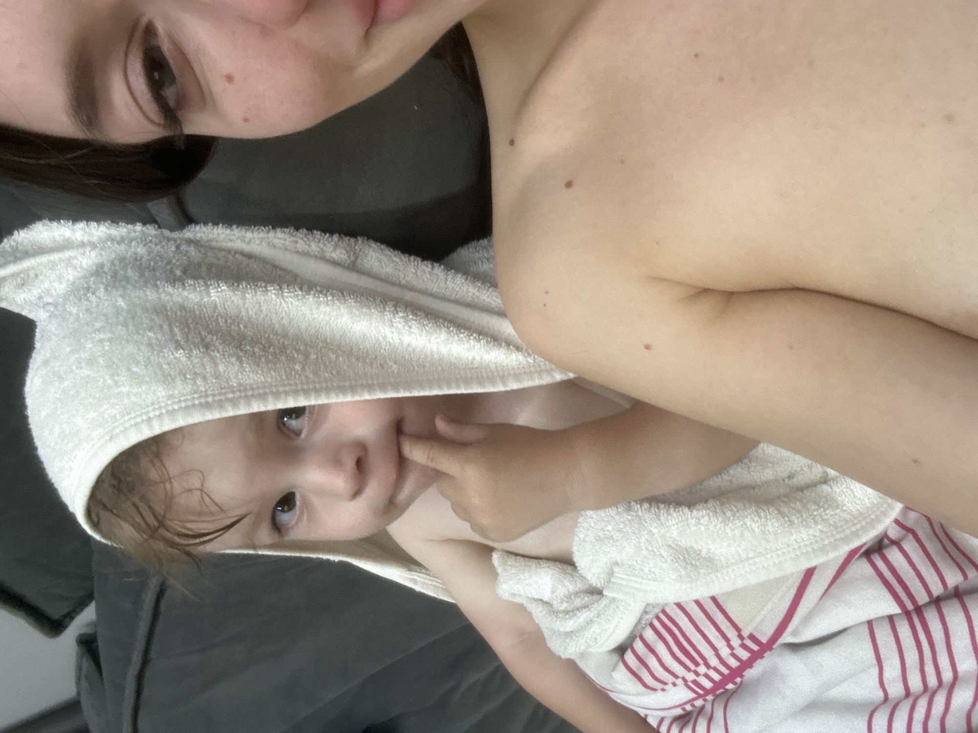 A close-up, shoulder-level selfie captures a woman on the right and a toddler on the left, both seemingly at home in Эстачи, Russia. The woman, with her face partially visible and her shoulder exposed, is looking into the camera with a slight smile. The toddler, wrapped in a white hooded towel with pink stripes underneath, is positioned to the left of the woman. The toddler's eyes are open, and they are looking to the side with one finger in their mouth. The background consists of a dark, plush surface, possibly a sofa. The lighting appears to be natural, creating a soft ambiance.
