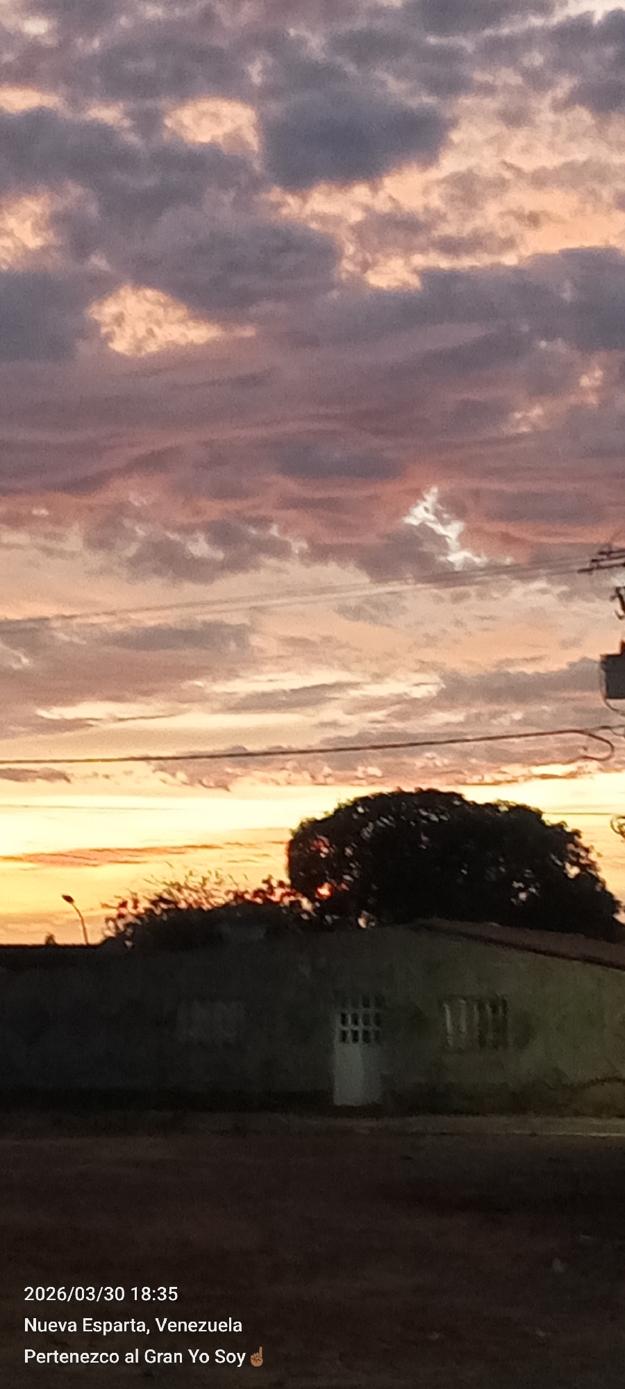 The image, taken at 18:35 on March 30, 2026, in Nueva Esparta, Venezuela, shows a building with a dark roof and a door and window. Above the building is a tree with dark foliage. In the background is a sunset with orange and pink hues in the sky, with dark clouds. Power lines are visible in the upper right portion of the image. The city of Altagracia is not mentioned.