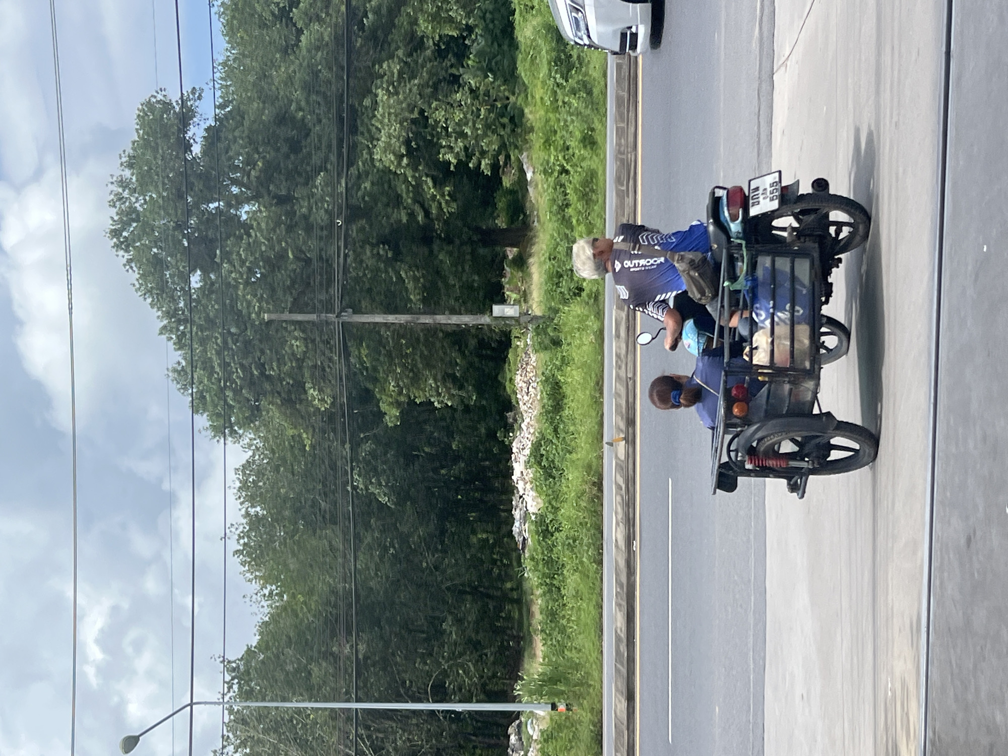 Two people are on a three-wheeled motorcycle with a sidecar. The driver, a person with white hair and sunglasses, wears a blue and black cycling jersey. A passenger with dark hair pulled into a ponytail, wearing a blue shirt, sits in the sidecar. The motorcycle has a license plate with Thai characters and the number "555". They are on a paved road next to a grassy median with trees and a pile of debris in the background. A white car is partially visible on the right. The scene is in Pa Khlok, Thailand, with a lamppost and power lines overhead.