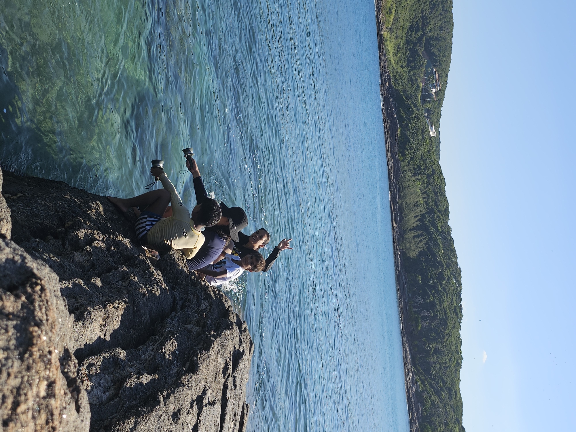 Four individuals are positioned on a rocky outcrop adjacent to the ocean. The rocky surface is dark in color. The ocean exhibits a turquoise tint near the rocks, transitioning to a deeper blue further out. One person is pointing to an object beyond the frame. A coastline covered in lush greenery stretches into the distance. Two people are holding cylinder objects. The location is Armação dos Búzios, Brazil.