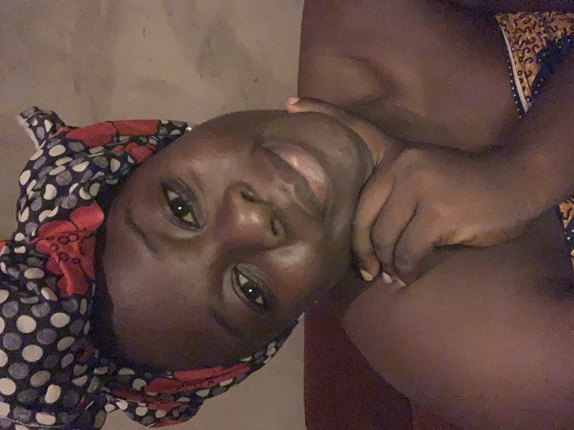 The image shows a close-up, slightly grainy, self-portrait of a Black woman wearing a headscarf. She is looking directly at the camera with her chin resting on her hand. Her headscarf has a dark blue, white, and red polka dot and floral pattern. She is wearing a patterned garment around her shoulders and upper chest, with a colorful band visible at the bottom of the frame. The background appears to be a plain, textured wall. The lighting is dim, creating a warm glow on her skin. The woman's features are prominent, with her eyes open and a slight smile on her lips. Her skin appears smooth and dewy. The overall impression is intimate and relaxed. The image was taken in Agban, Nigeria.