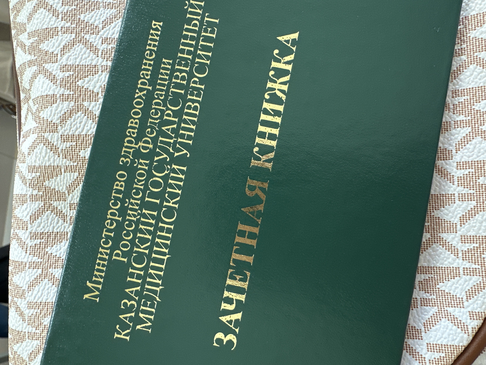 The image shows a dark green book, likely a student record book or "grade book," placed diagonally on a white and brown patterned surface. The pattern appears to be a repeating monogram, possibly a brand logo.
The book's cover features embossed gold text in Cyrillic. The text reads:
"Министерство здравоохранения
Российской Федерации
КАЗАНСКИЙ ГОСУДАРСТВЕННЫЙ
МЕДИЦИНСКИЙ УНИВЕРСИТЕТ"
Which translates to:
"Ministry of Healthcare
of the Russian Federation
KAZAN STATE
MEDICAL UNIVERSITY"
Below this, larger gold text reads:
"ЗАЧЕТНАЯ КНИЖКА"
Translating to:
"GRADE BOOK" or "RECORD BOOK."
The close-up shot focuses primarily on the book and the immediate background, suggesting an indoor setting. There is no human activity visible. The text explicitly links the object to Kazan, Russia, aligning with the provided location context. The lighting suggests ambient indoor illumination, with no specific time of day indicated.