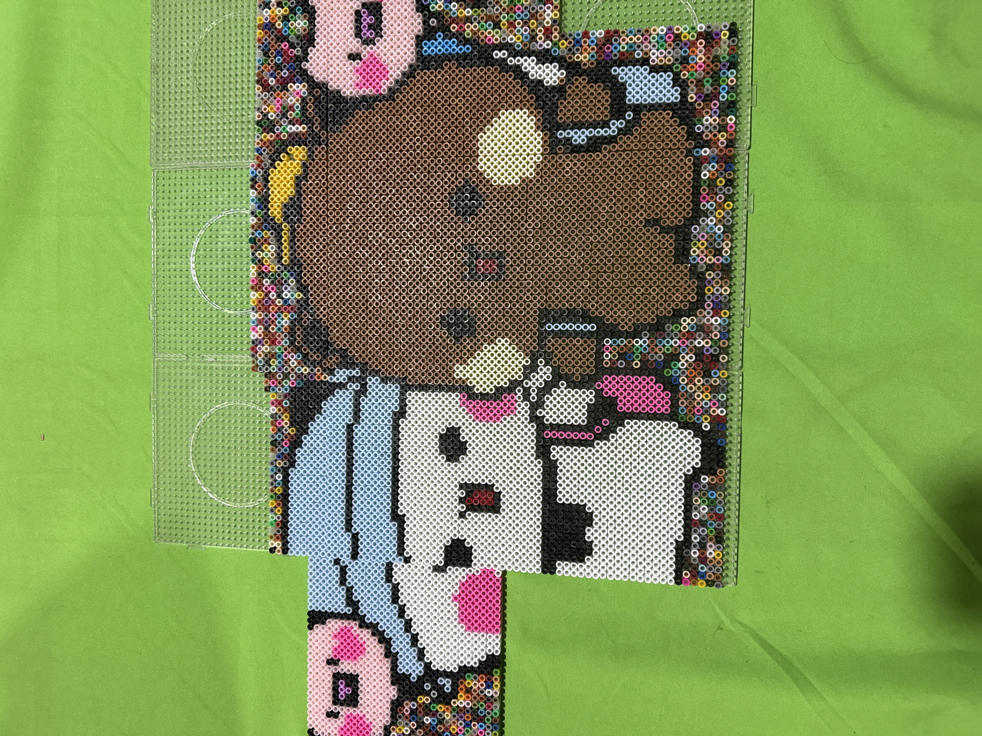 The image depicts a partially completed Perler bead artwork laid on a green fabric surface. The artwork is a pixelated representation of a cartoon character, likely a dog or bear, wearing a blue outfit and holding a white object. The character's head is brown with a yellow circular highlight. The border surrounding the character is made of a variety of colorful beads. To the left of the artwork is a clear plastic Perler bead grid, possibly used as a template for the design. The image was taken indoors, with the green fabric acting as a backdrop. The location within Puebla, Mexico, is not evident from the image's content.
