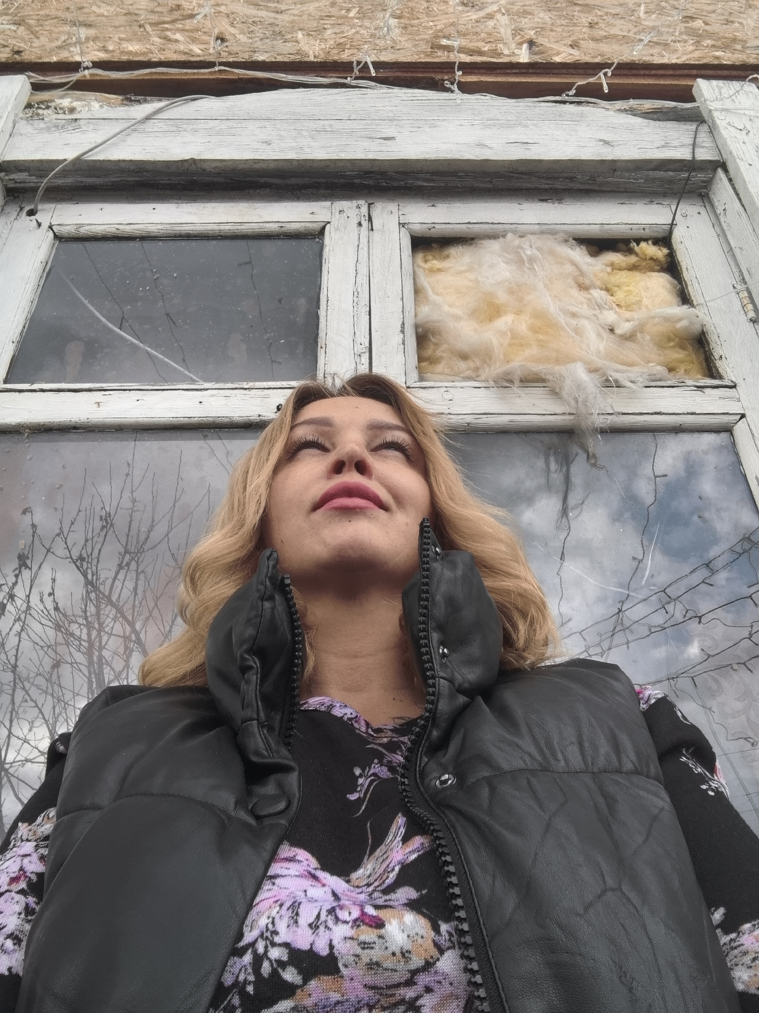 A low-angle shot features a blonde woman in a black vest looking upward in front of an old window. The window frame is painted white and appears weathered, with peeling paint. The panes are cracked or broken, and insulation is visible in one of the window openings. The reflection in the remaining glass panes suggests a cloudy sky and bare tree branches. The woman's clothing consists of a floral-patterned shirt beneath the black vest. The environment looks run-down, possibly abandoned. The city of Noviy, Russia is not discernible from the image alone.