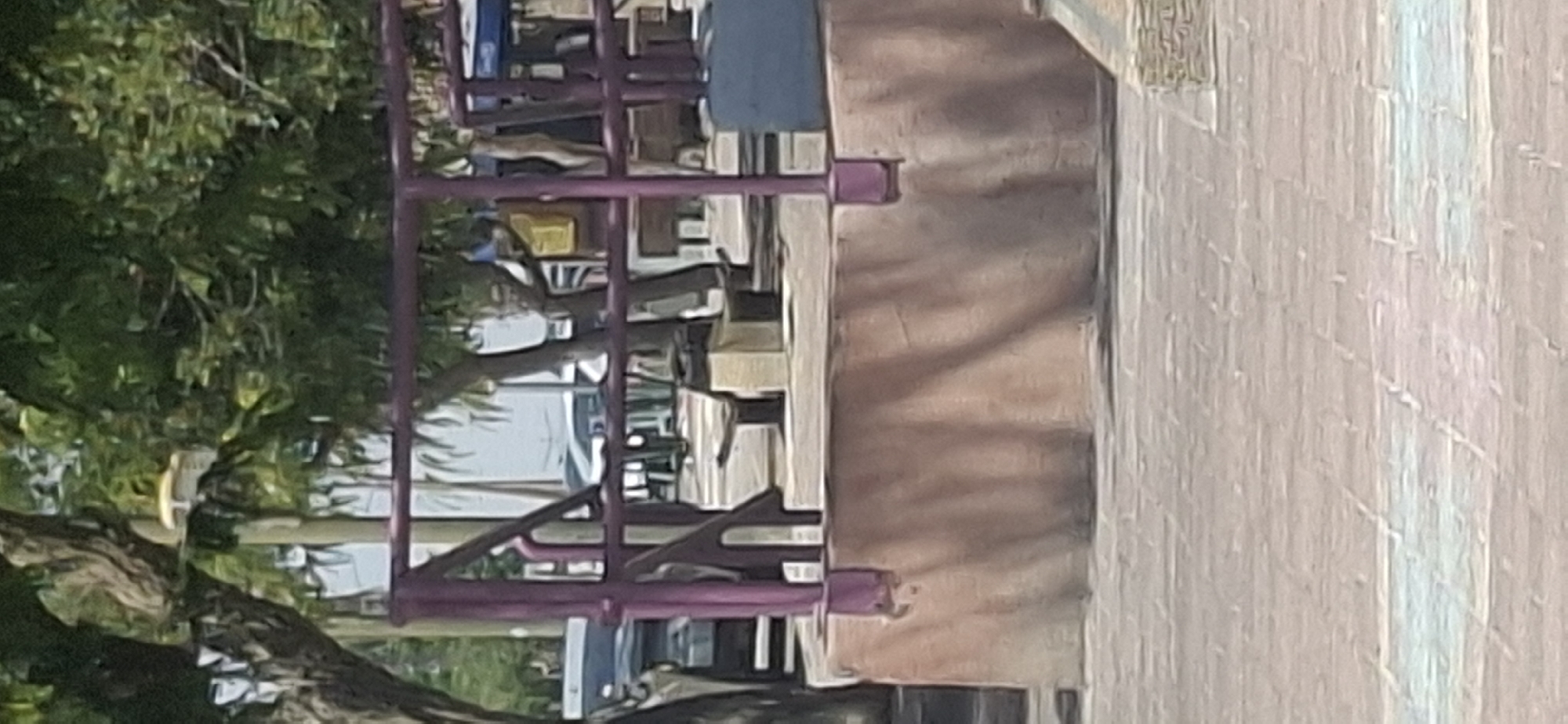 The image shows a public seating area with a purple railing in the foreground. Behind the railing, there are two concrete benches with wooden slats. A tree with green leaves is visible in the background, along with other trees and some cars. The ground is paved with interlocking bricks. The scene appears to be an outdoor recreational space, possibly a park or a plaza. There are no people or animals visible in the image. The overall impression is of a quiet, unoccupied public area. This scene is consistent with the public spaces found in the city of Yerocham, Israel, known for its parks and recreational areas.