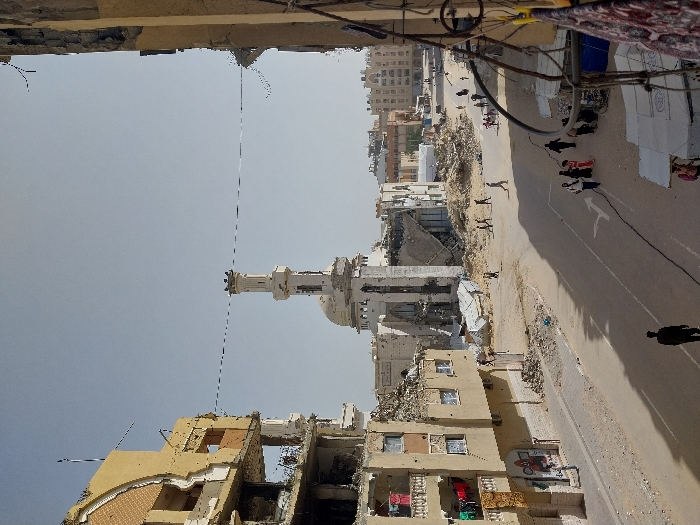 The image depicts a heavily damaged urban street scene in Khan Yunis, Palestinian Territory, under a clear, bright sky. In the midground, a large mosque structure stands prominently, with its dome and surrounding buildings significantly collapsed and reduced to rubble, though its central minaret remains largely intact. To the left, a multi-story building exhibits extensive structural damage, with outer walls collapsed, exposing multiple floors and internal rooms; laundry hangs from balconies on its lower intact sections. To the right, another partially visible building also shows damage.
The foreground features a paved road, partially covered with sand and debris, on which multiple individuals, including men, women, and children, are walking. Several groups of pedestrians are visible on the street and sidewalks, some navigating around rubble piles. On the immediate right, makeshift shelters or stalls with blue tarpaulins and fabric are set up. Extensive piles of concrete, twisted metal, and other building debris are scattered throughout the scene, particularly around the base of the mosque and the damaged buildings. Overhead, several thin wires or cables are strung across the street between structures. Road markings, including a white directional arrow, are visible on the asphalt. The overall environment conveys widespread destruction and debris, with ongoing civilian presence.