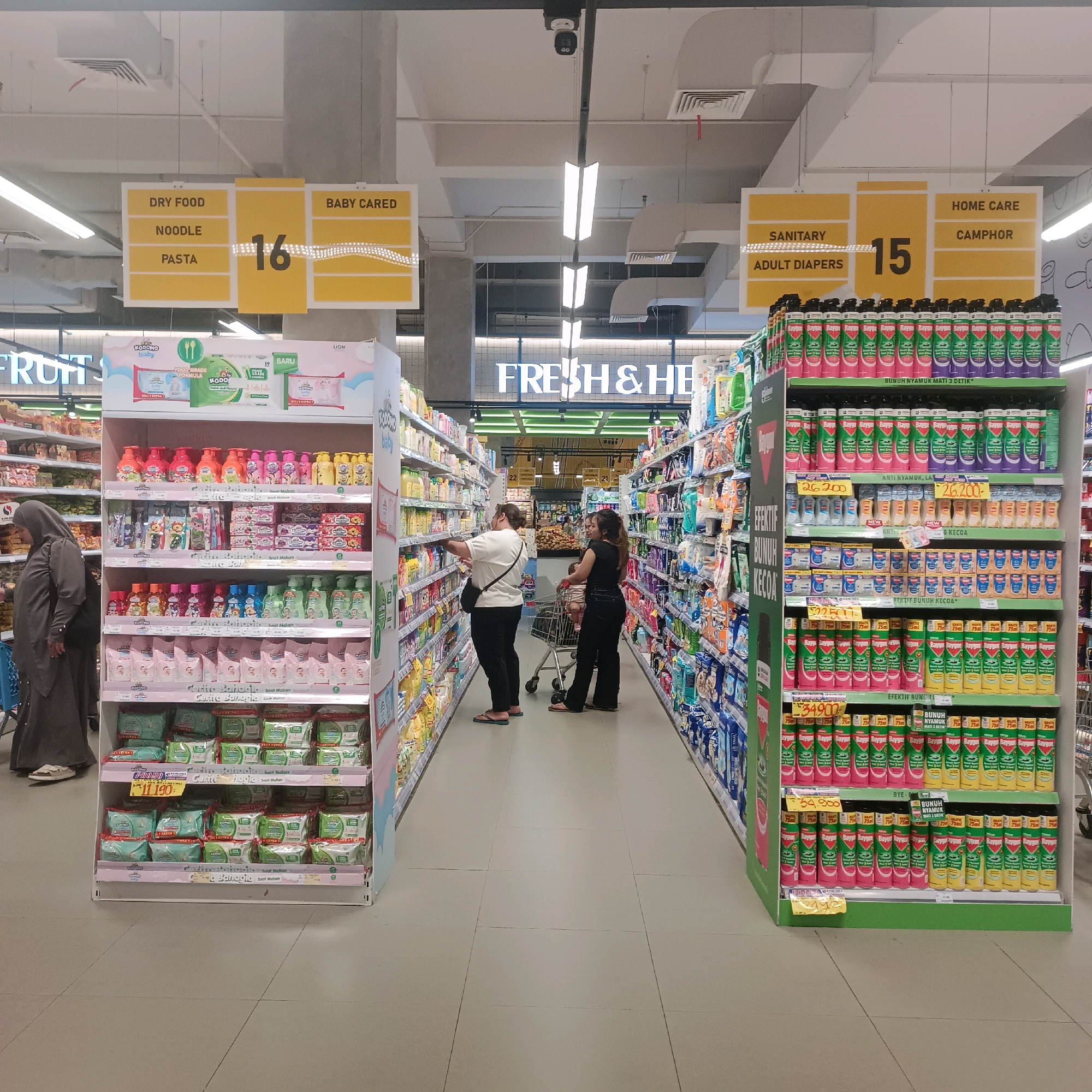 In a brightly lit supermarket aisle, shoppers navigate between shelves stocked with various products. Signs overhead indicate different sections of the store, such as "DRY FOOD," "NOODLE," "PASTA," "BABY CARED," "SANITARY," "ADULT DIAPERS," "HOME CARE," and "CAMPHOR." In the foreground, a woman wearing a grey hijab is browsing baby products on a display shelf. Further down the aisle, two women are pushing a shopping cart, with one examining items on a shelf. The shelves are filled with colorful packaging, including baby care items, cleaning supplies, and what appear to be food products. The floor is tiled with a light gray pattern. The overall impression is a typical supermarket scene in Surabaya, Indonesia, with an emphasis on hygiene and family-oriented products.