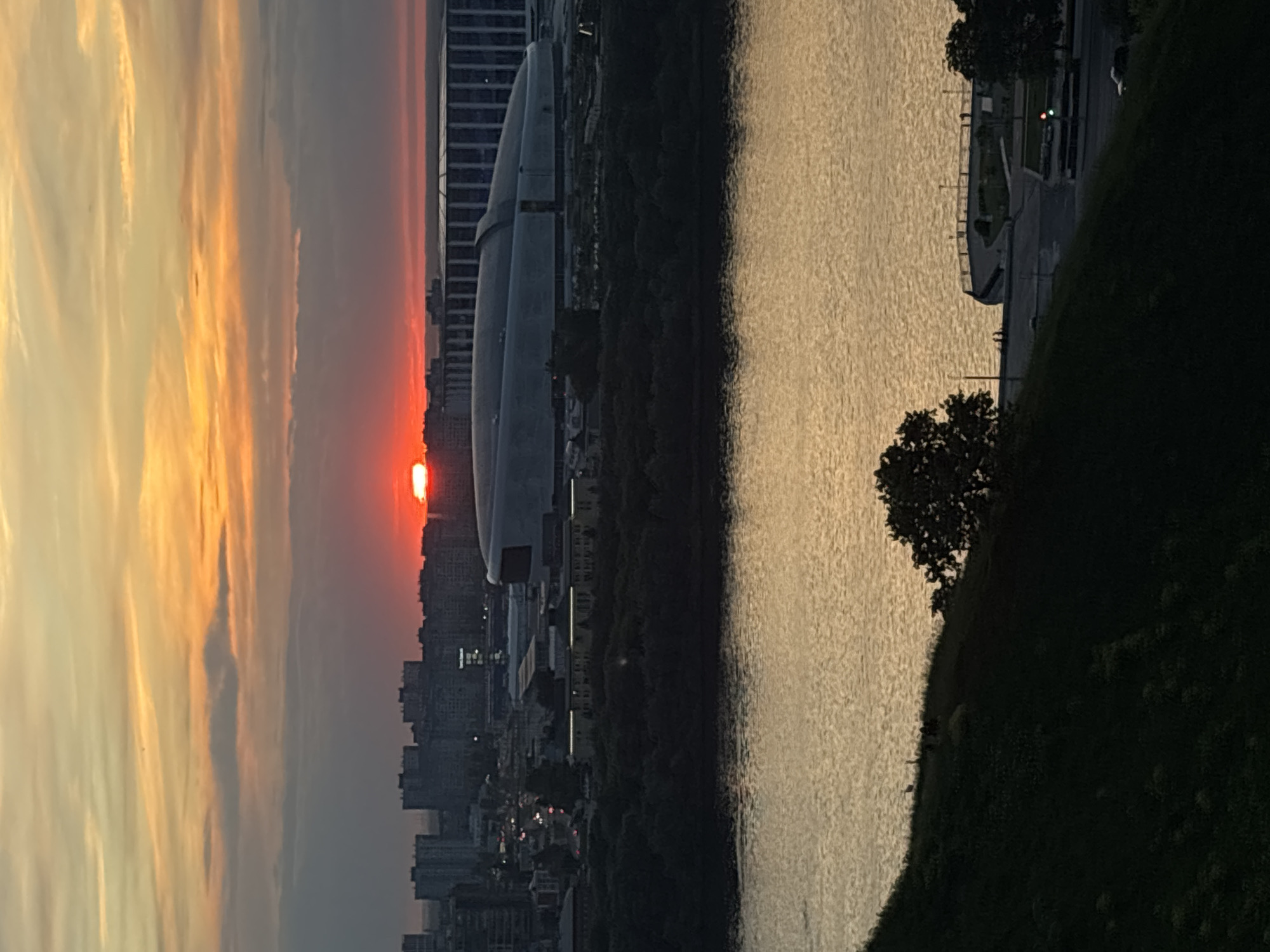The image captures a vibrant sunset over a city and a wide river, viewed from an elevated vantage point, likely overlooking Nizhny Novgorod, Russia. The sun, a brilliant orange-red disc, is setting low on the horizon, partially obscured by the distant urban skyline. The sky is filled with sweeping clouds, illuminated in fiery shades of orange, red, and yellow near the horizon, gradually fading to softer grays and blues higher up.
Across the mid-ground, a broad river stretches, its surface shimmering with reflections of the twilight sky, appearing as a textured expanse of muted golden-brown. On the far bank of the river, a dense cityscape is silhouetted against the colorful sky. Notable among the buildings is a distinctive modern structure with a large, curved roof. As dusk settles, faint lights are beginning to twinkle within some of the city buildings.
In the immediate foreground, a dark, grassy hill or bank slopes downwards towards the river on the right side of the frame. Scattered trees, their forms dark against the brighter water, are visible on this bank. Along the riverside closer to the foreground, a paved area or walkway is discernible, featuring railings and a visible green traffic light, suggesting a road or pedestrian area.
The activity is the natural progression of a sunset. The scene is calm and serene, with no identifiable people or explicit events taking place. There is no visible text in the image.