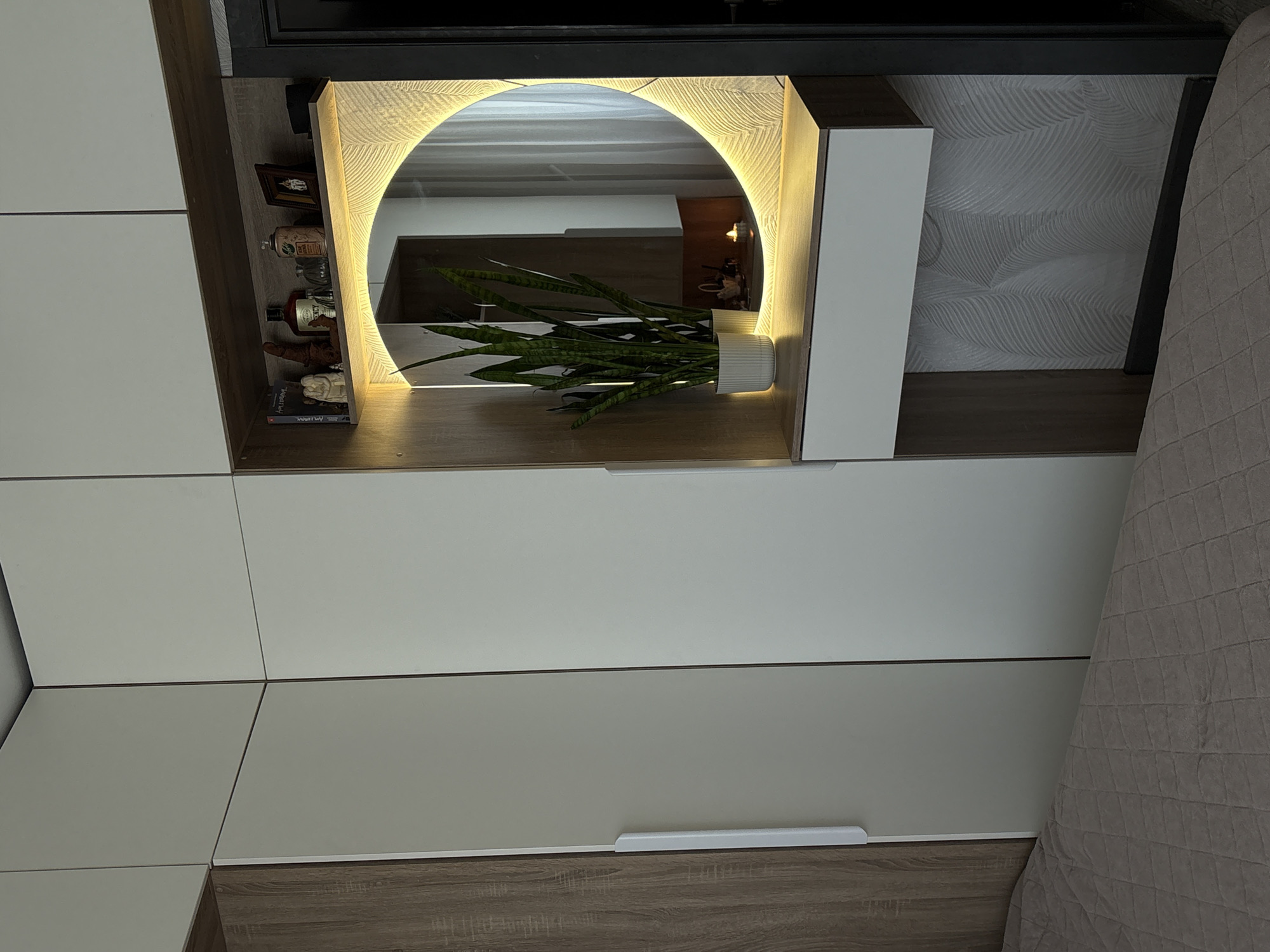 The image shows a modern interior design detail. A built-in shelf unit displays a circular, backlit mirror. A snake plant in a white pot sits in front of the mirror. The shelf unit incorporates white cabinets and drawers with wood trim. The unit also features decorative items on a shelf to the left of the mirror. A bed with a quilted cover is partially visible to the right of the shelf unit. The scene does not provide enough information to determine whether it is located in Birobidzhan, Russia.