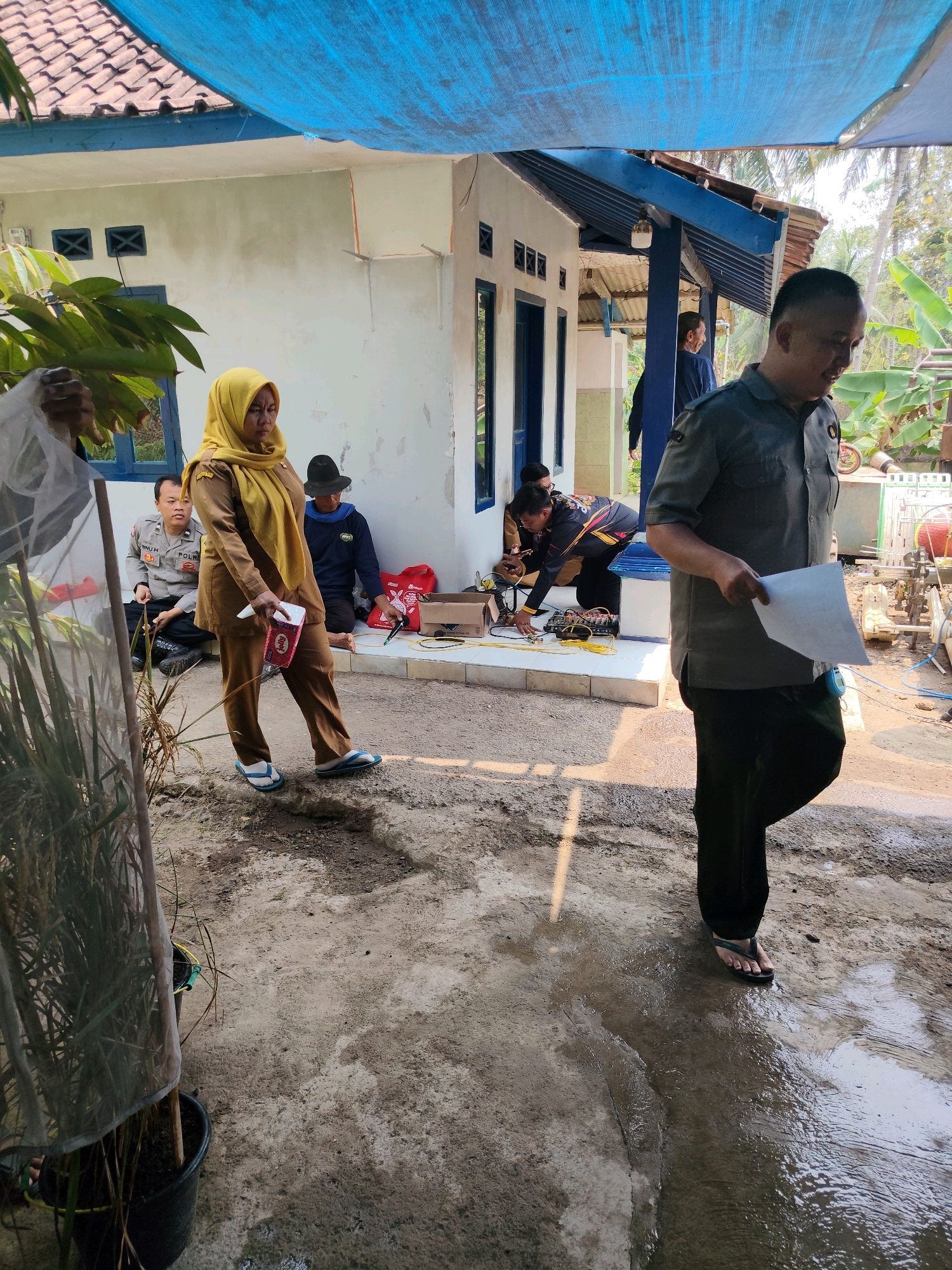 In Karyamukti, Indonesia, a group of individuals are gathered outdoors in front of a building. A woman in a yellow hijab and a brown uniform stands holding a box of tissues. To her left, a man in a police uniform is seated. In the background, several other people are present, some appearing to be working with electrical equipment. The ground is a mixture of dirt and puddles, with some vegetation visible. The scene suggests an outdoor event or work activity taking place on a sunny day.