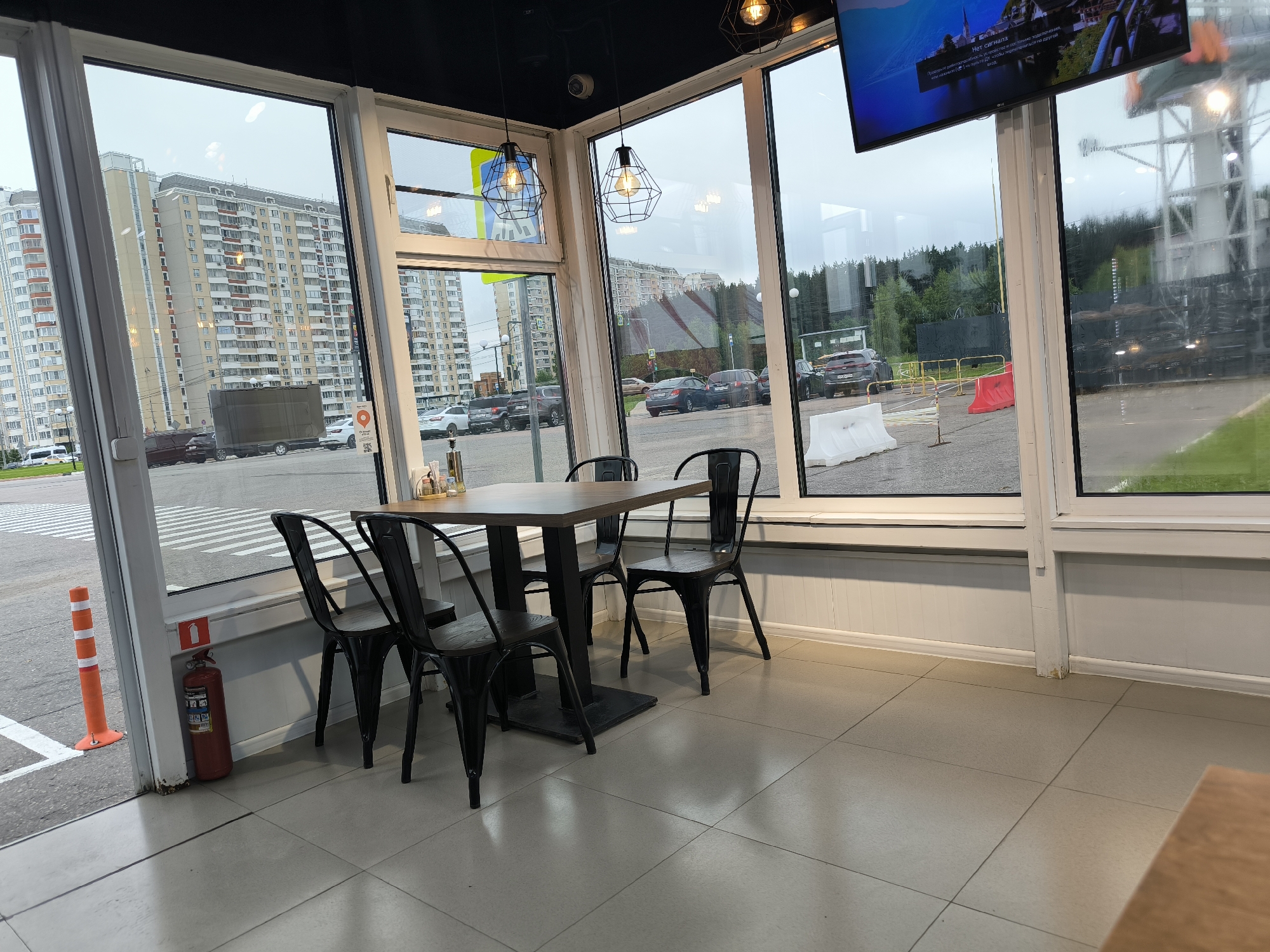 The image captures an indoor view from a cafe or restaurant in Lobnya, Russia, looking out through large windows onto a street and surrounding buildings. Inside, a dark wooden table with black metal chairs is set up. The interior features light-colored tiled flooring and white wainscoting. A red fire extinguisher is visible near the entrance. Outside, a street with pedestrian crossings, parked cars, and tall residential buildings can be seen. A CCTV camera is mounted on the ceiling inside. The sky appears overcast.