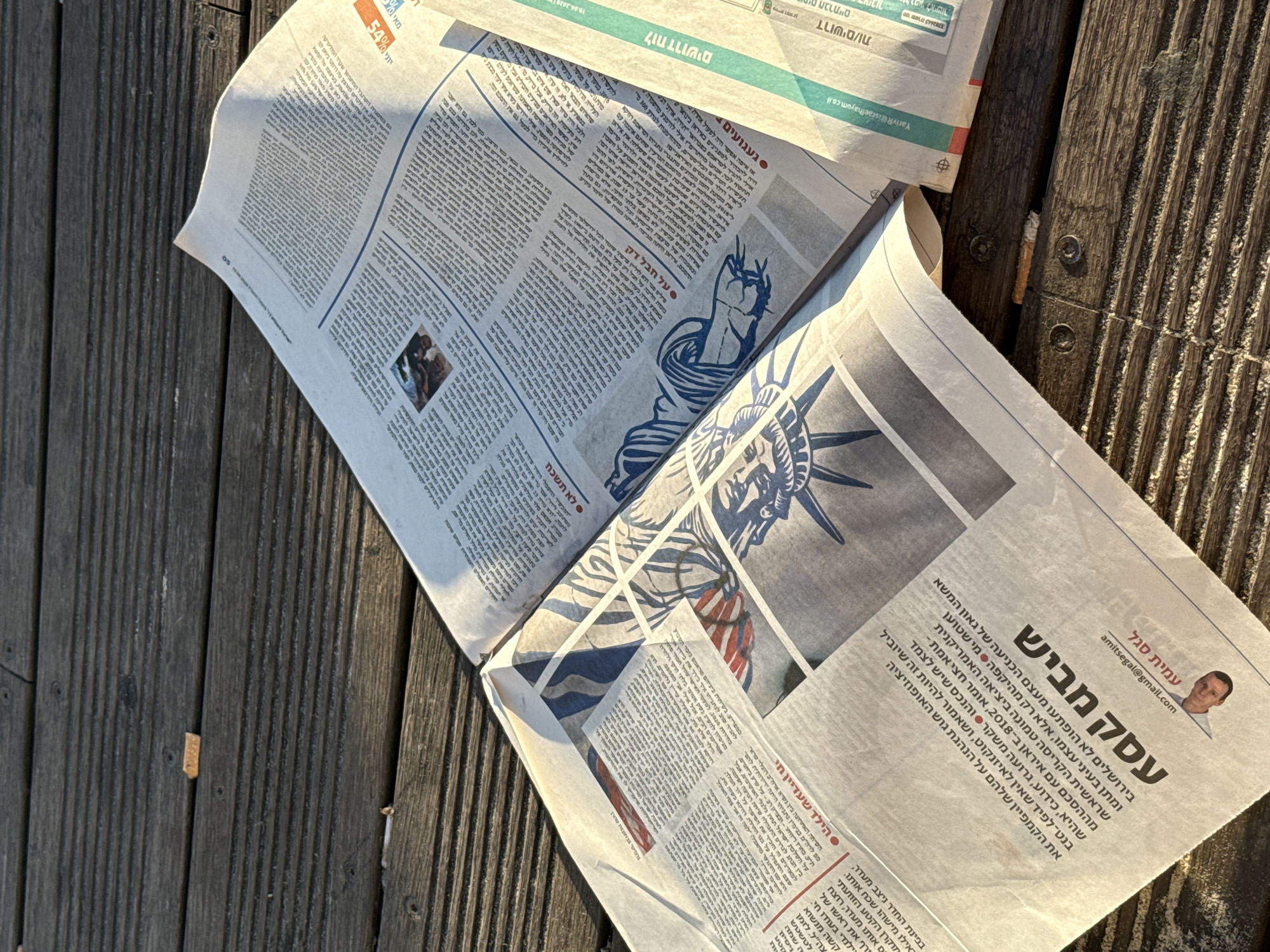 An open newspaper lies on a weathered wooden surface, possibly a deck or boardwalk. The newspaper is printed in Hebrew and features an illustration of the Statue of Liberty. The lighting suggests it is late afternoon or early morning, casting long shadows. The wooden surface is made of planks with visible grain and some small debris, such as cigarette butts, are present. The image appears to be taken in Tel Aviv, Israel, given the presence of Hebrew text. There are no people visible in the image.