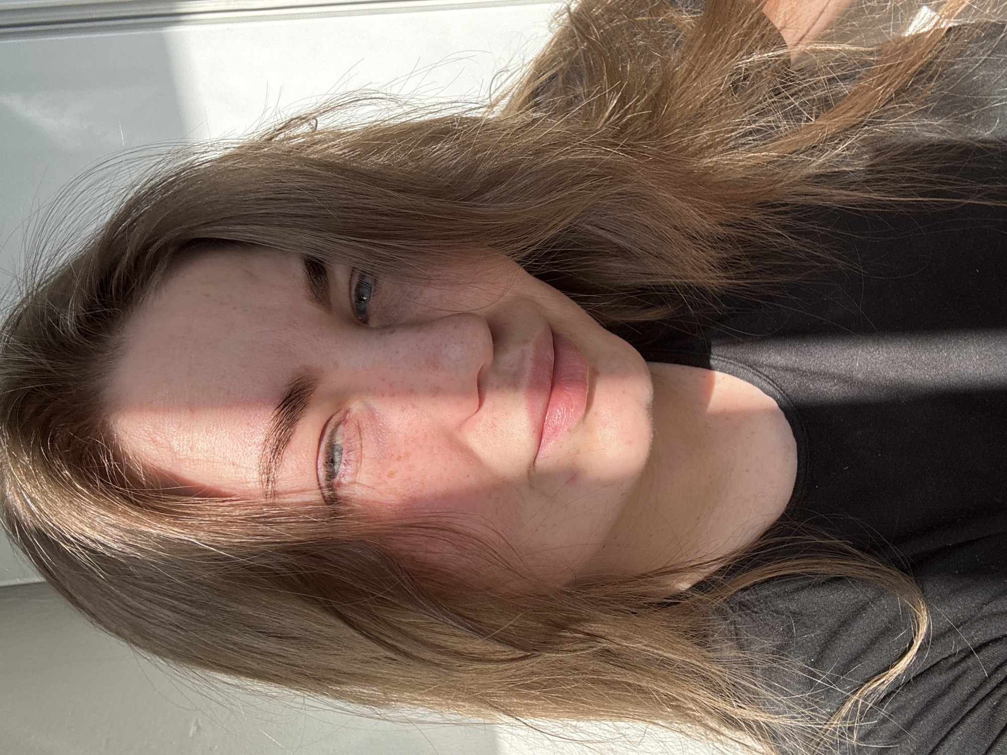 A close-up portrait captures a person, likely a young woman, lying down and looking directly towards the viewer. The image is rotated 90 degrees counter-clockwise. She has long, flowing light brown hair that cascades around her face and extends over a plain black top she is wearing. Her eyes are a light blue or grey, and her face features fair skin with numerous freckles visible on her nose and cheeks. Her lips are pink and slightly parted, contributing to a neutral or softly pleasant expression.
The scene is illuminated by strong, direct sunlight, which casts distinct shadows. A sharp line of shadow diagonally crosses her face and upper body, leaving one side brightly lit and the other in shade. This natural lighting highlights the texture and color of her hair. The background is simple and out of focus, appearing as a bright, uniform surface behind her head, possibly a wall or bedding, and a darker area where her body rests.
The person appears to be posing for a photograph. The bright sunlight indicates it is daytime. There are no specific location cues, but the lighting suggests an indoor setting with natural light entering from a window. No visible text is present in the image.