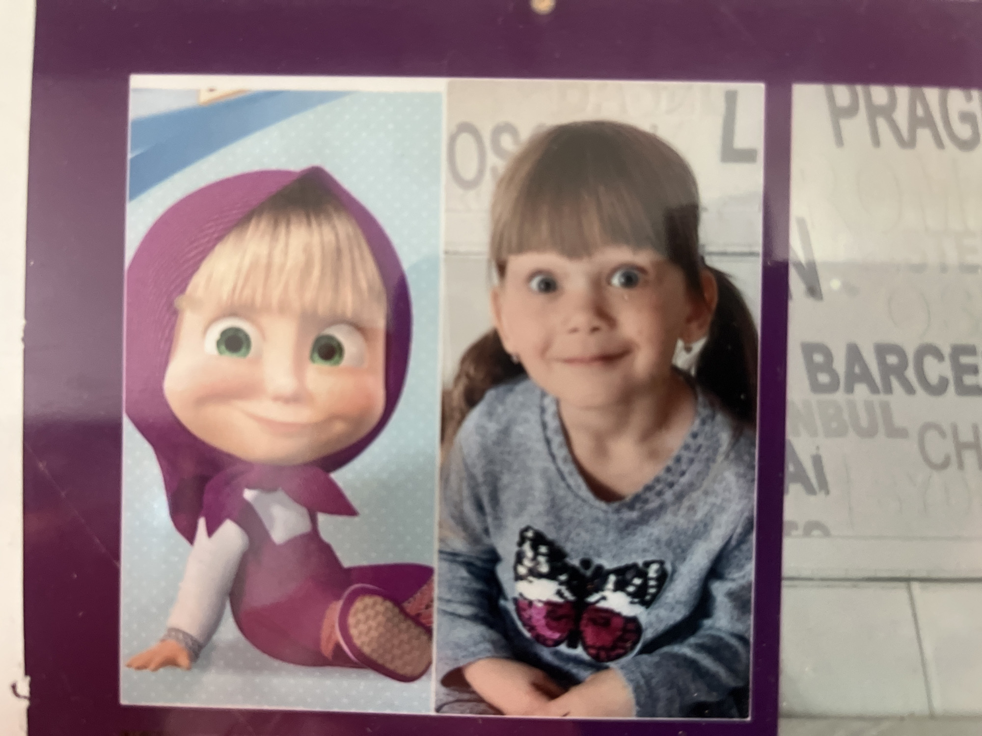 The image is a photograph of a young girl next to a cartoon character, likely from the animated show "Masha and the Bear." The girl has light brown hair styled with bangs and pigtails. She is wearing a gray long-sleeved shirt with a sequined butterfly design on the front. Her expression is wide-eyed and slightly surprised. The cartoon character is Masha, depicted in her signature purple outfit with a matching headscarf. She has a smiling expression and green eyes. The photograph appears to be a collage, with a purple border framing the two images. The background behind the girl includes blurred text, suggesting it might be a collage of travel-related images or souvenirs. One section of the text clearly shows "PRAG" and "BARCE," likely referring to cities like Prague and Barcelona. The overall presentation suggests a personal memento, possibly displayed in a home or a souvenir shop. The image does not provide any specific visual information to determine its location within the city of Schwäbisch Hall, Germany.