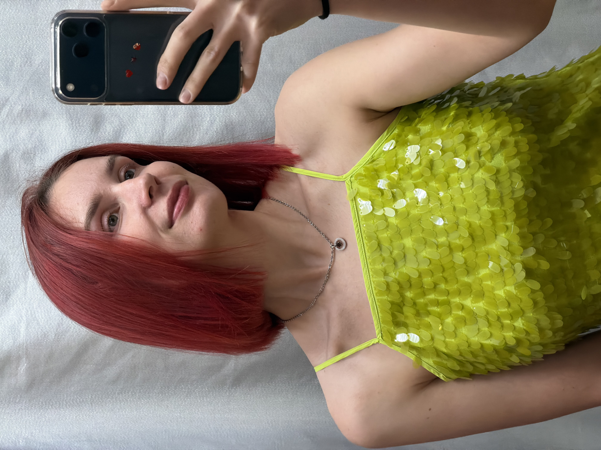 A woman with auburn hair is holding a smartphone, presumably taking a selfie. She is wearing a lime green, sequined top and a silver necklace. The background appears to be a white sheet. The location is not discernible from the image.