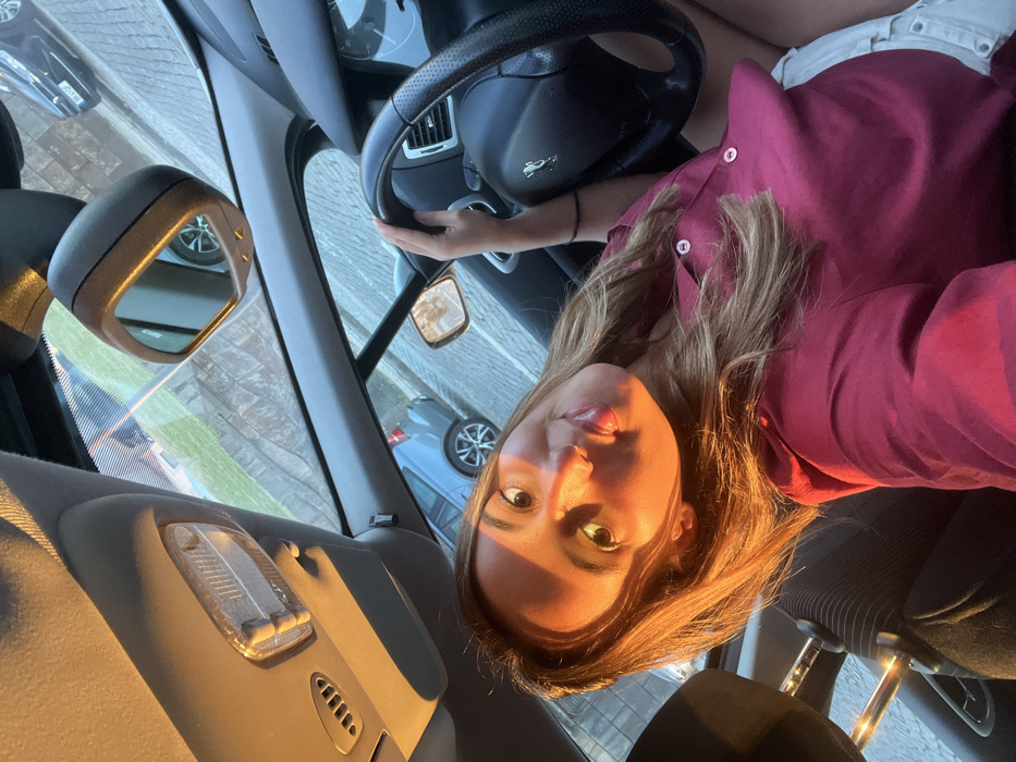 The image captures a young woman seated in the driver's seat of a car, illuminated by warm, golden sunlight characteristic of late afternoon or early evening. She has long, light brown hair that catches the light and is wearing a maroon or dark pink collared shirt. Her face is turned towards the camera, displaying a calm expression and a slight smile, with her eyes looking forward.
Her right hand rests on the steering wheel, which prominently features a Peugeot lion emblem. The car's interior is visible, including parts of the dashboard, a rearview mirror, and an overhead interior light fixture. Through the front windshield and reflected in the side mirror, other cars are visible on a paved surface, suggesting the car is parked on a street or in a parking lot. The rearview mirror also shows reflections of another vehicle and a building.
The scene conveys a sense of peacefulness, highlighted by the soft, directional sunlight casting warm tones on the woman's face and hair. The setting is identified as being within Aldoar, Foz do Douro e Nevogilde, Portugal.