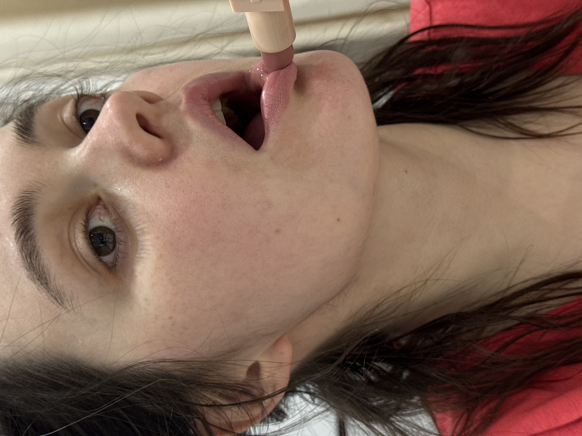 A person is lying on a light-colored surface with their head tilted back, applying light pink lipstick to their lower lip. The person has dark hair and is wearing a red shirt. The individual is facing the camera, and their mouth is slightly open, revealing their tongue. The image does not show any identifiable elements that would indicate the location of Izhevsk, Russia.